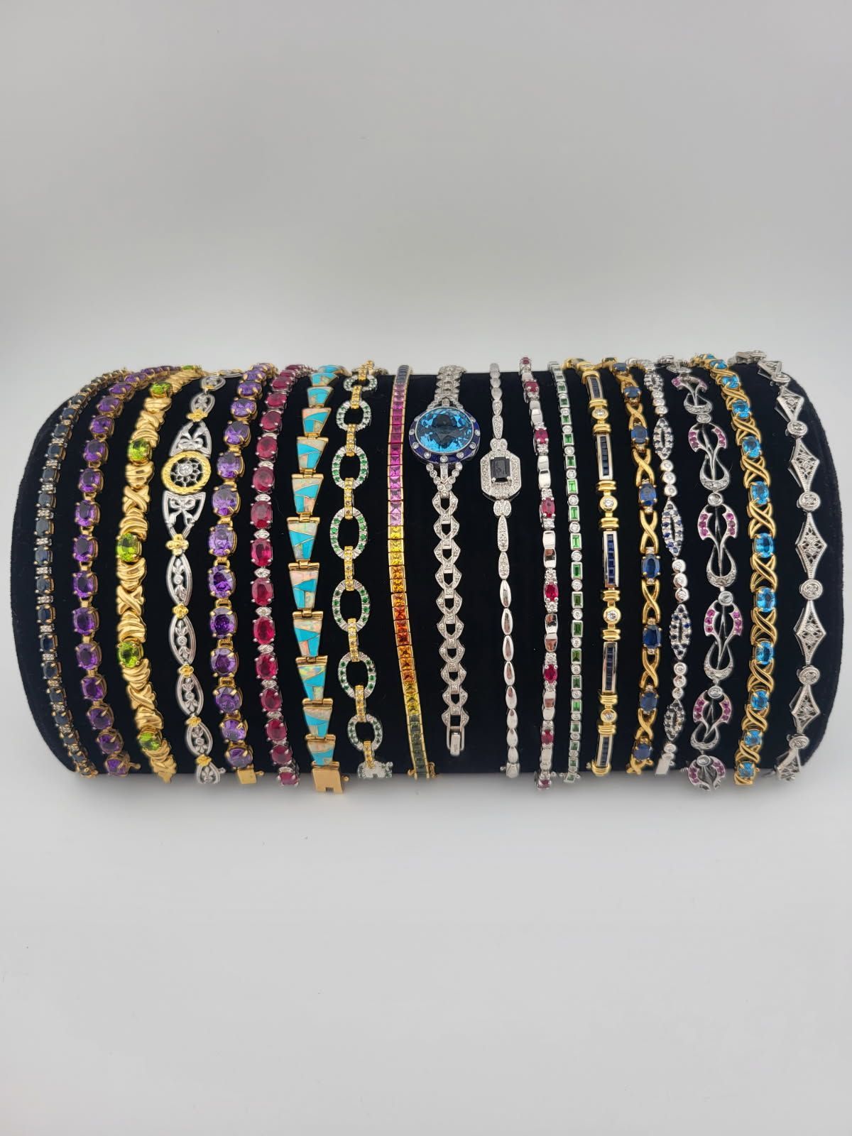 Assortment of gem-studded bracelets displayed on a black cylindrical stand against a light gray background.