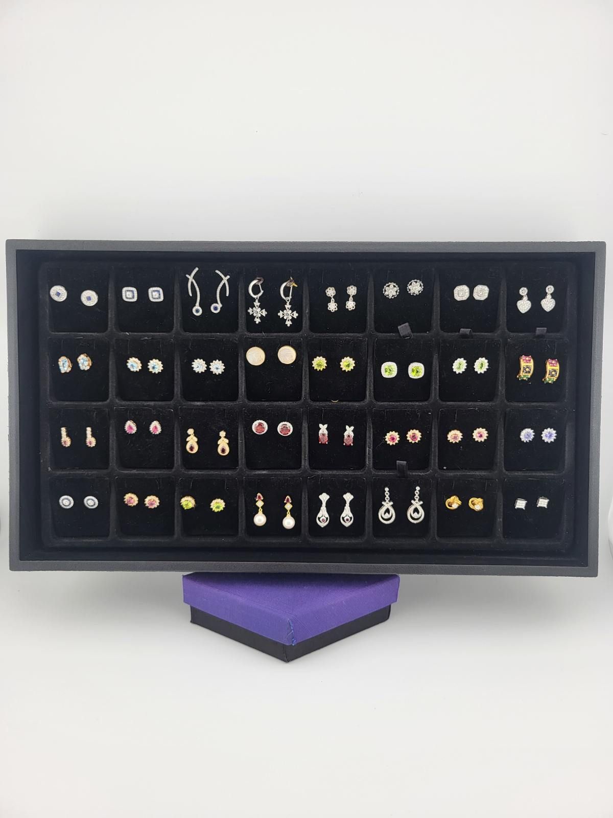 A black jewelry display case filled with stud earrings of various designs and colors. A purple box is below.