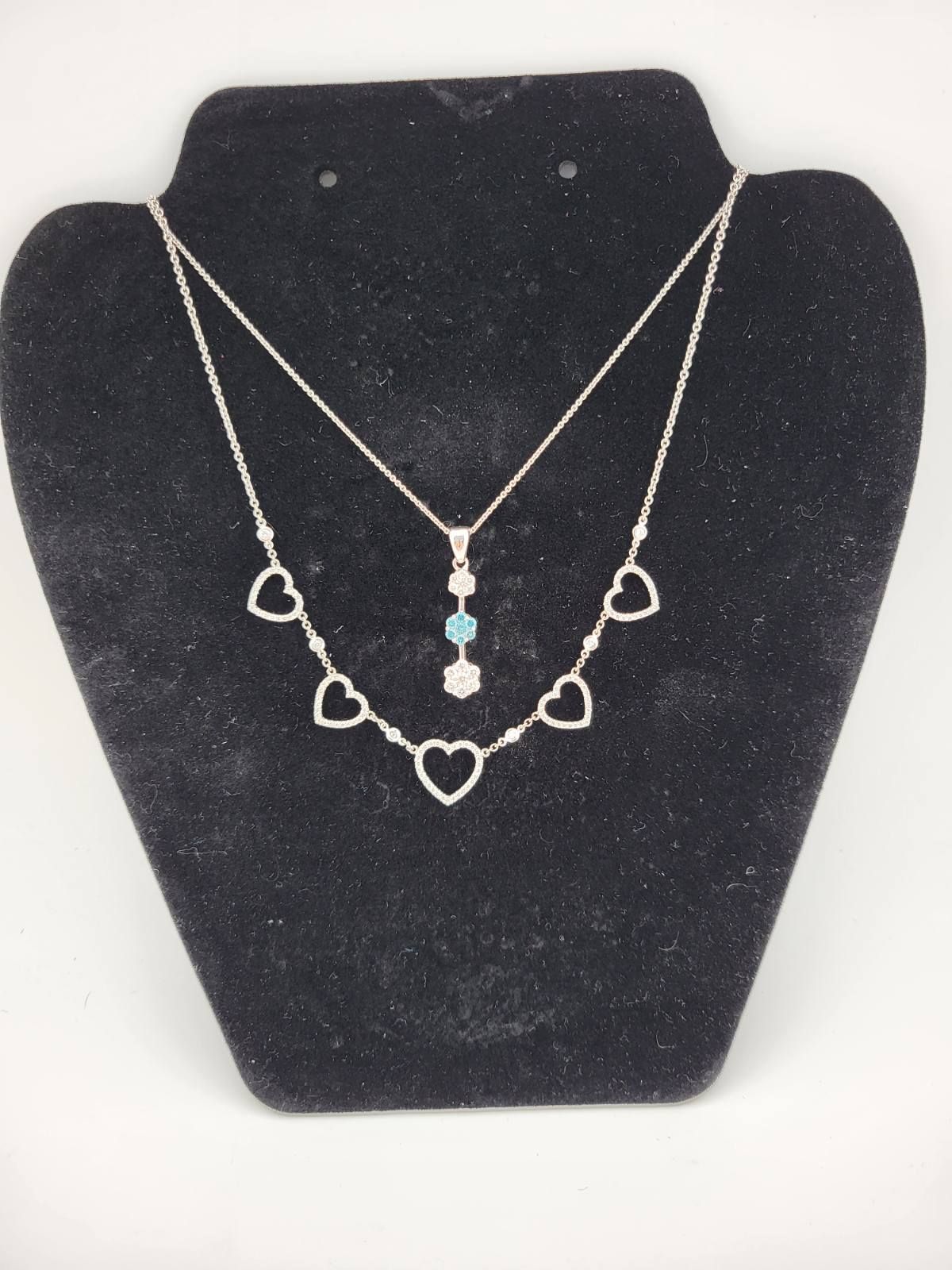 Two silver necklaces on a black jewelry display. One has heart-shaped pendants; the other has a blue and white gem pendant.