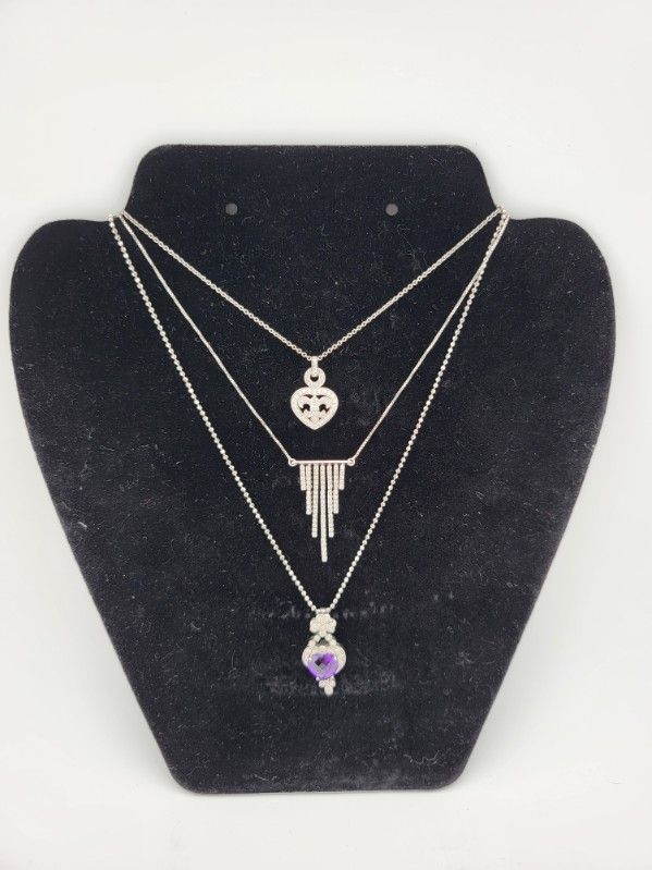 Three layered necklaces on a black jewelry display; silver chains, heart, geometric and amethyst pendant.