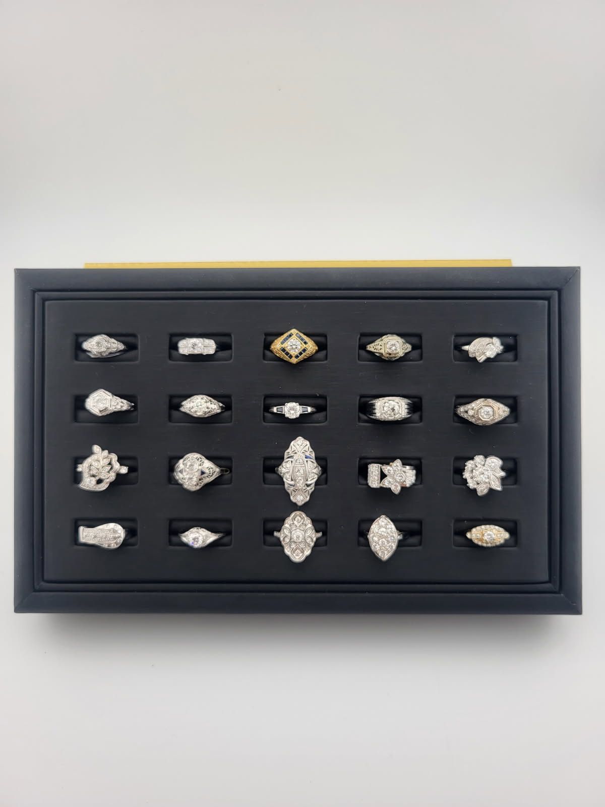 Black display tray holding twenty-five diamond rings. Several shapes, all silver or gold settings, against a white background.