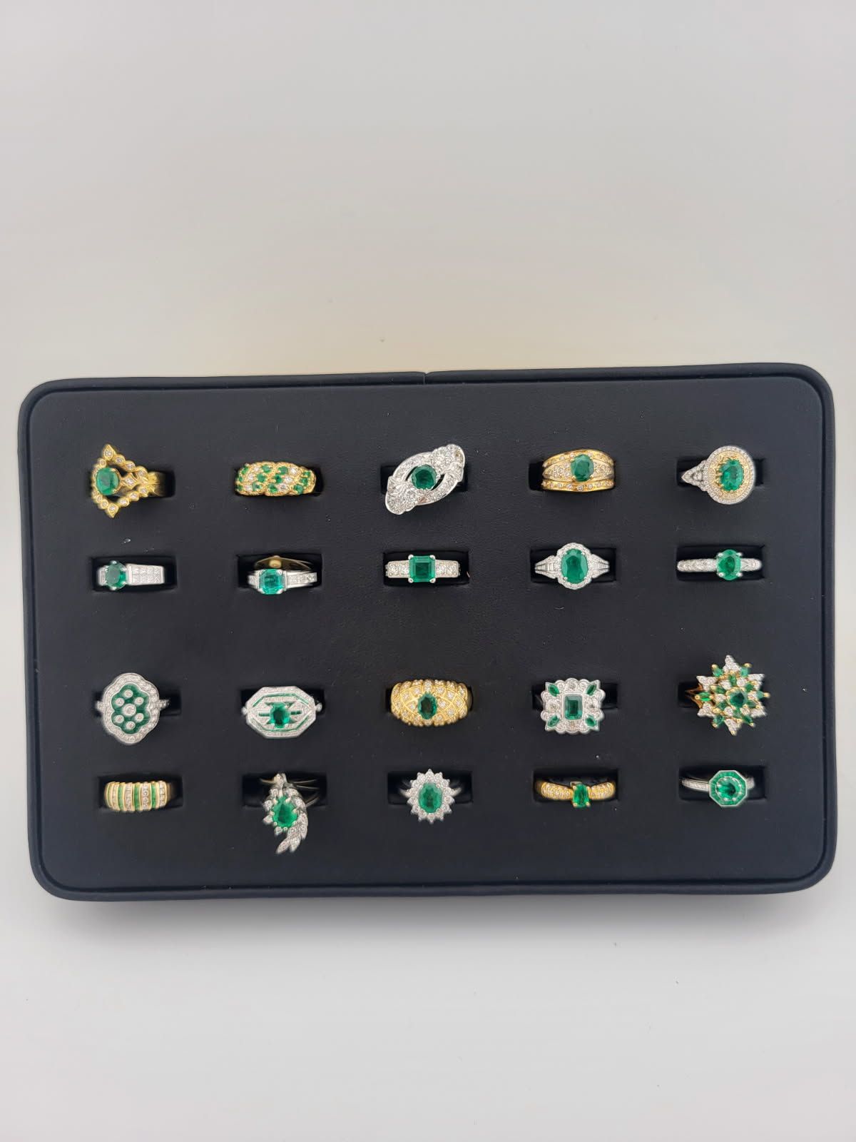 Black tray displaying twenty-four emerald gemstone rings of various designs.