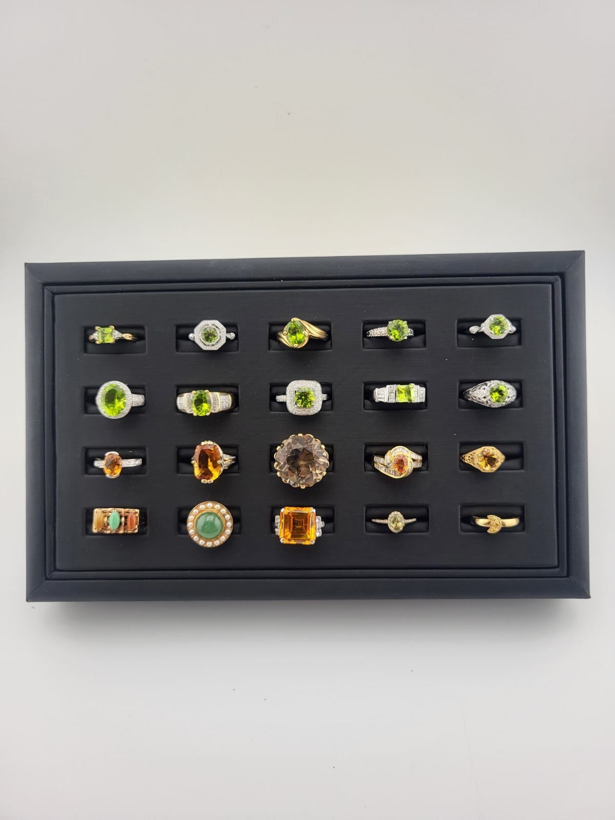 Black jewelry display case with a grid of gemstone rings, mostly gold and green.