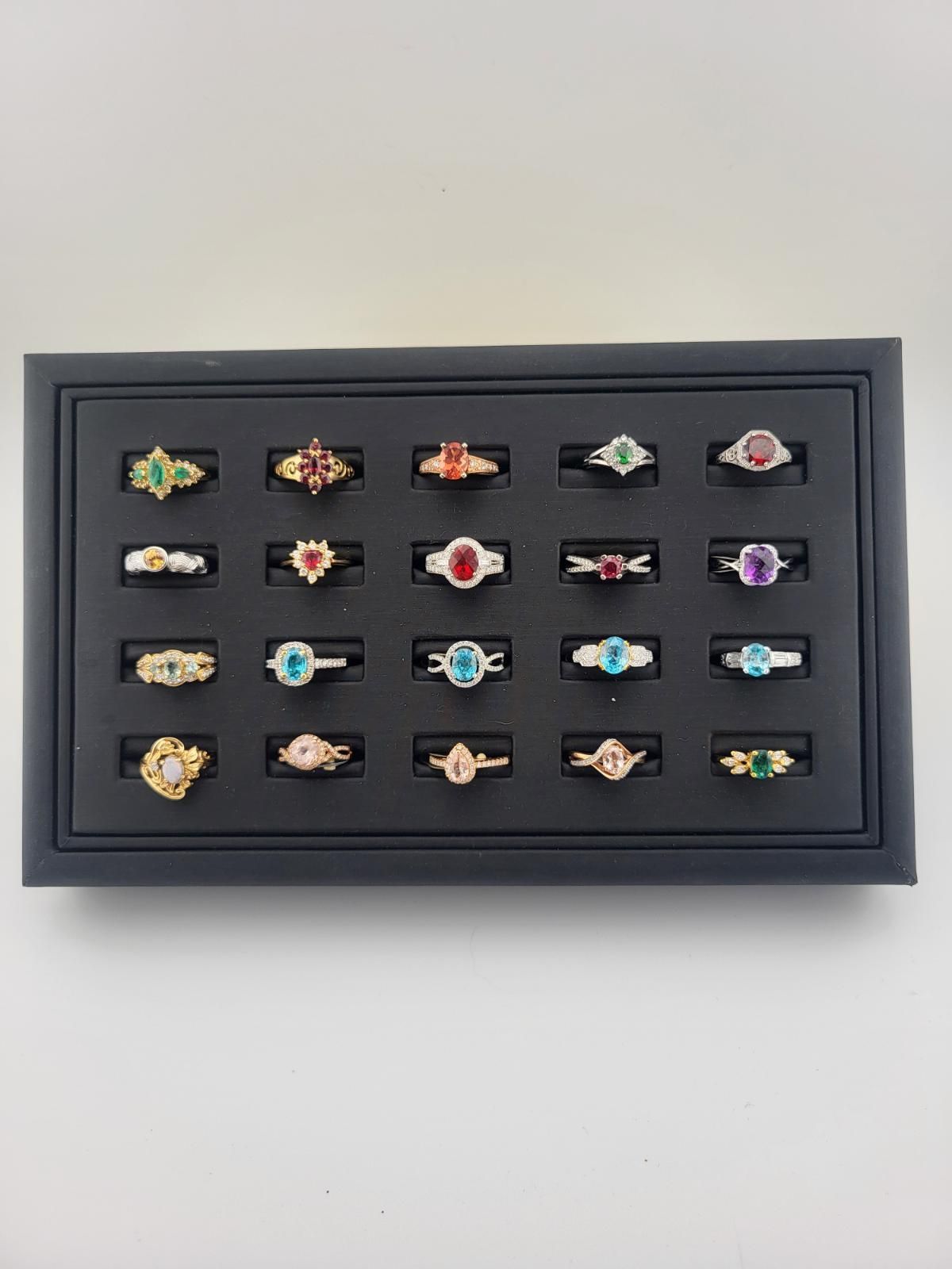 A display of twenty-four rings with various colored gemstones set in a black jewelry tray on a white surface.