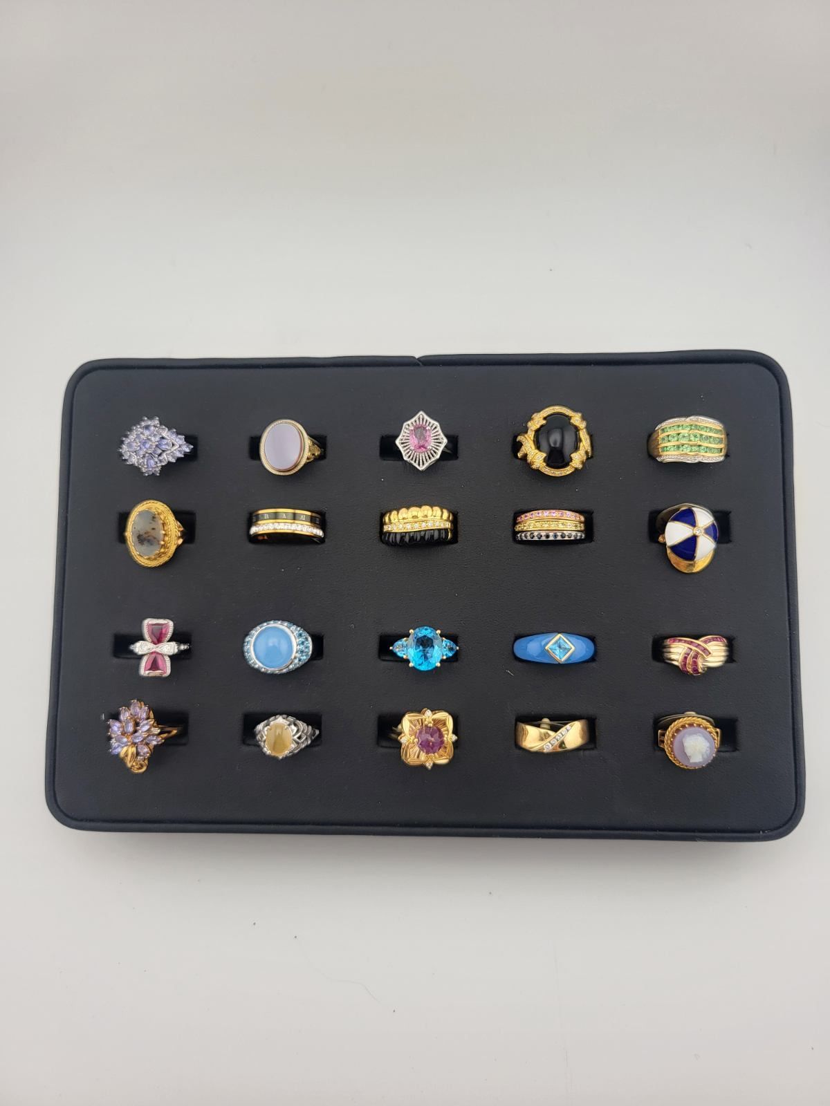 A tray of 24 colorful rings with various gemstones and designs on a black background.