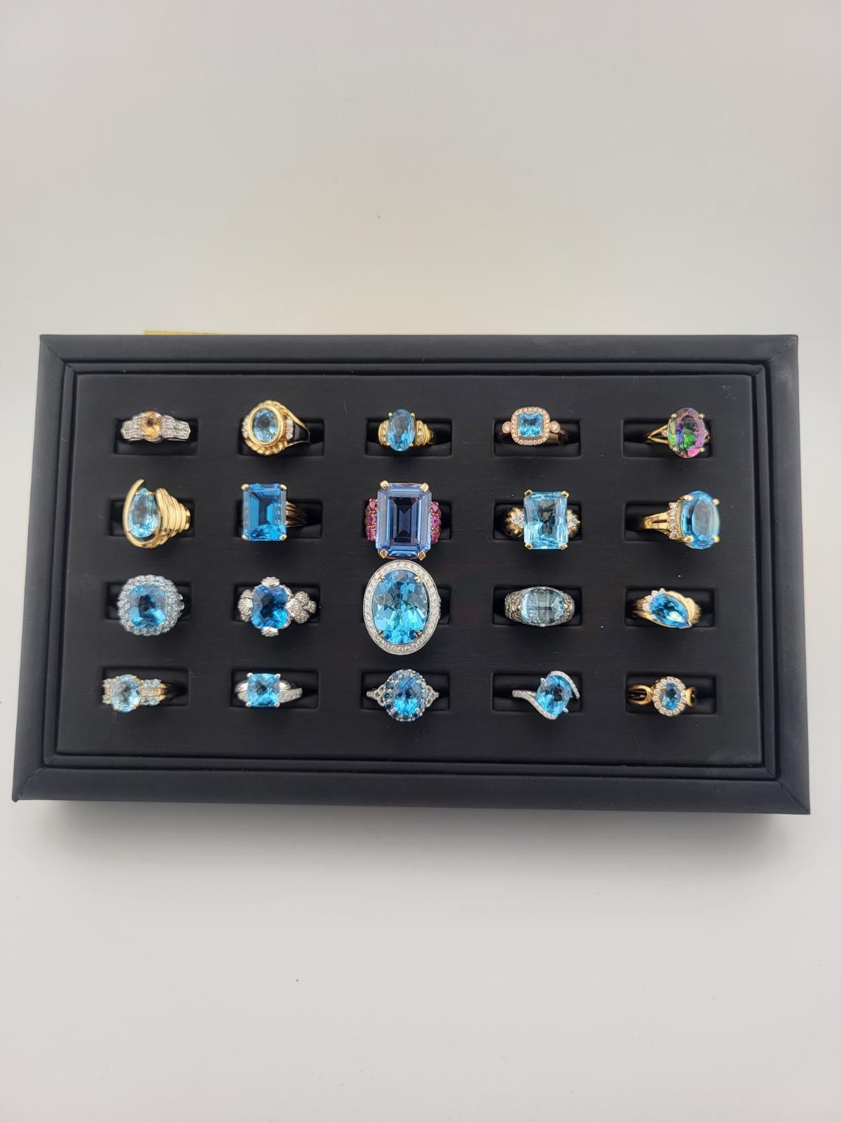 Black jewelry tray displaying a collection of blue topaz rings with gold and silver settings.
