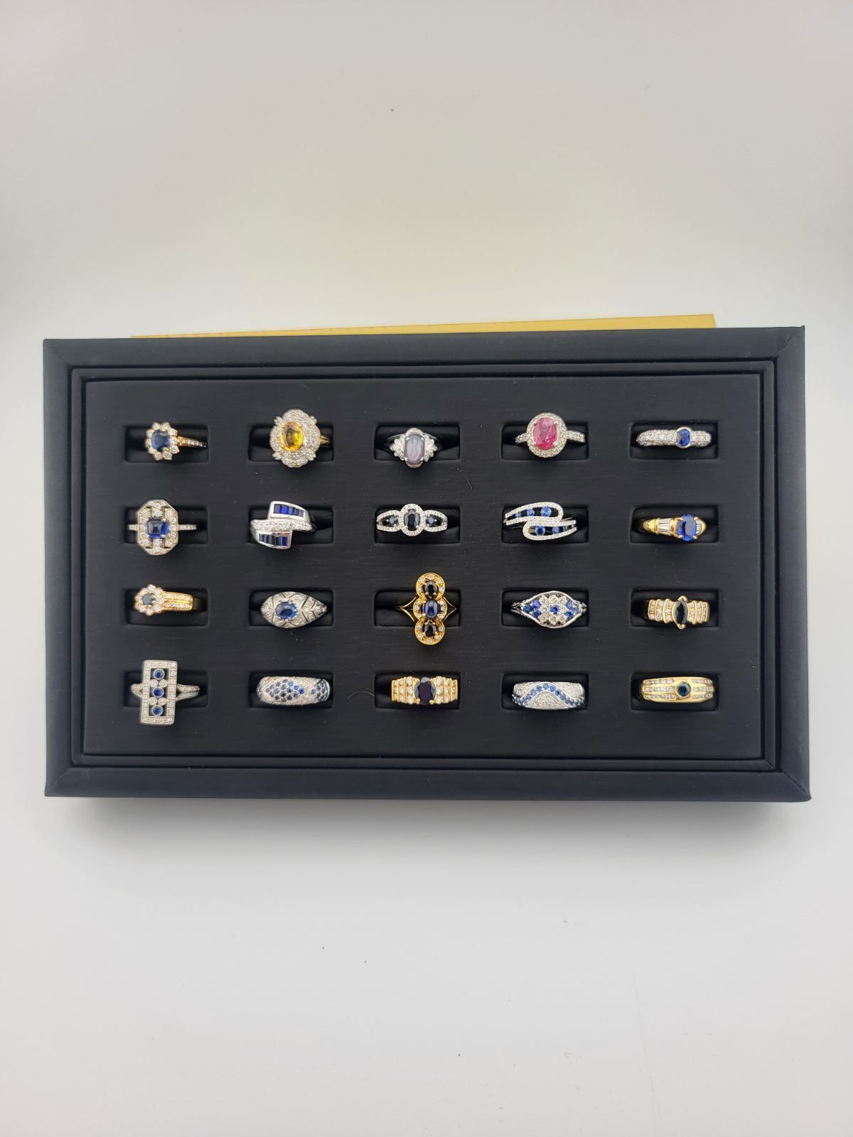 Black display tray with 24 assorted rings, mostly gold with colored gemstones, on a white background.
