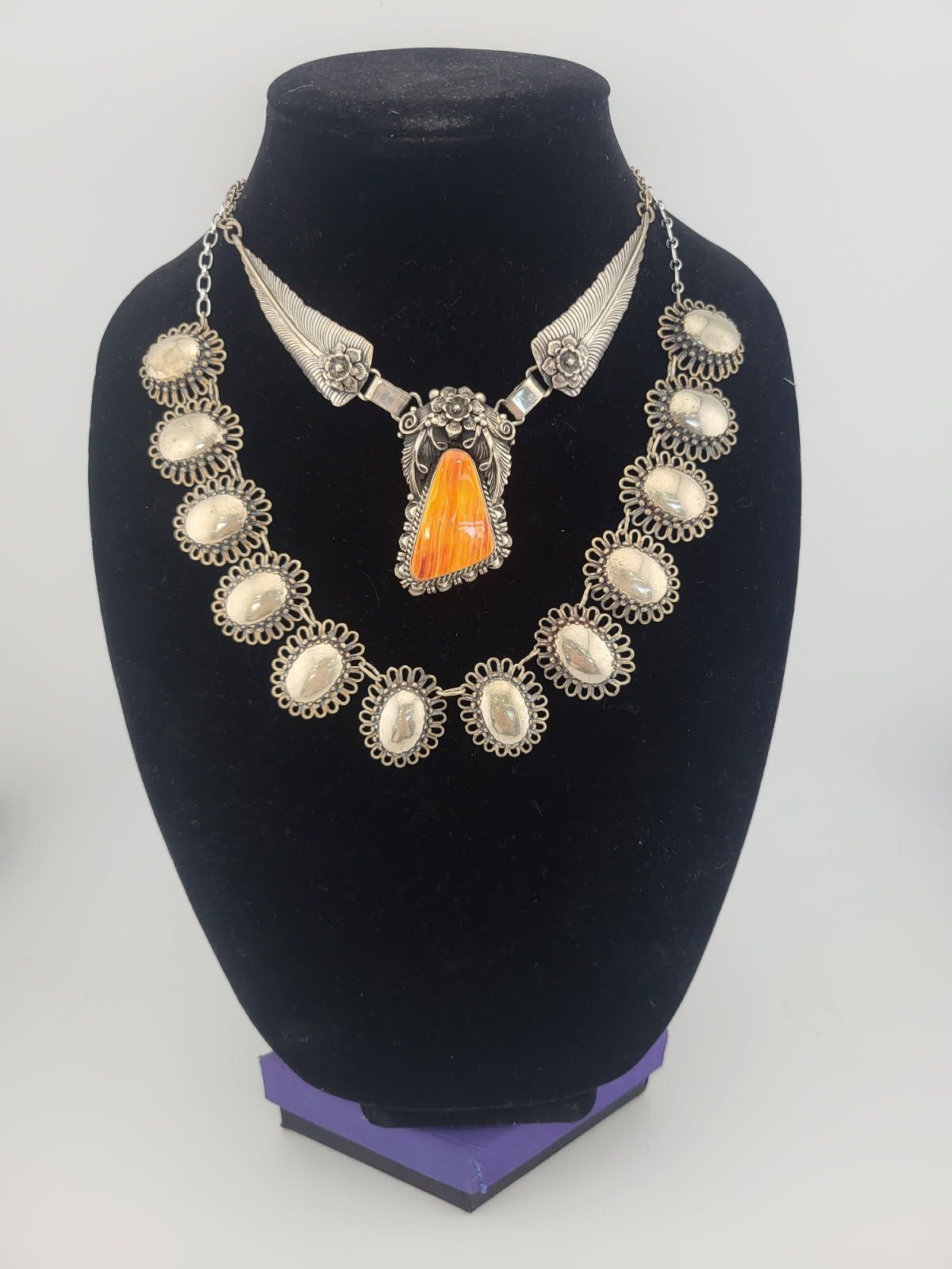 Two silver necklaces with oval embellishments and a central orange stone on a black display.
