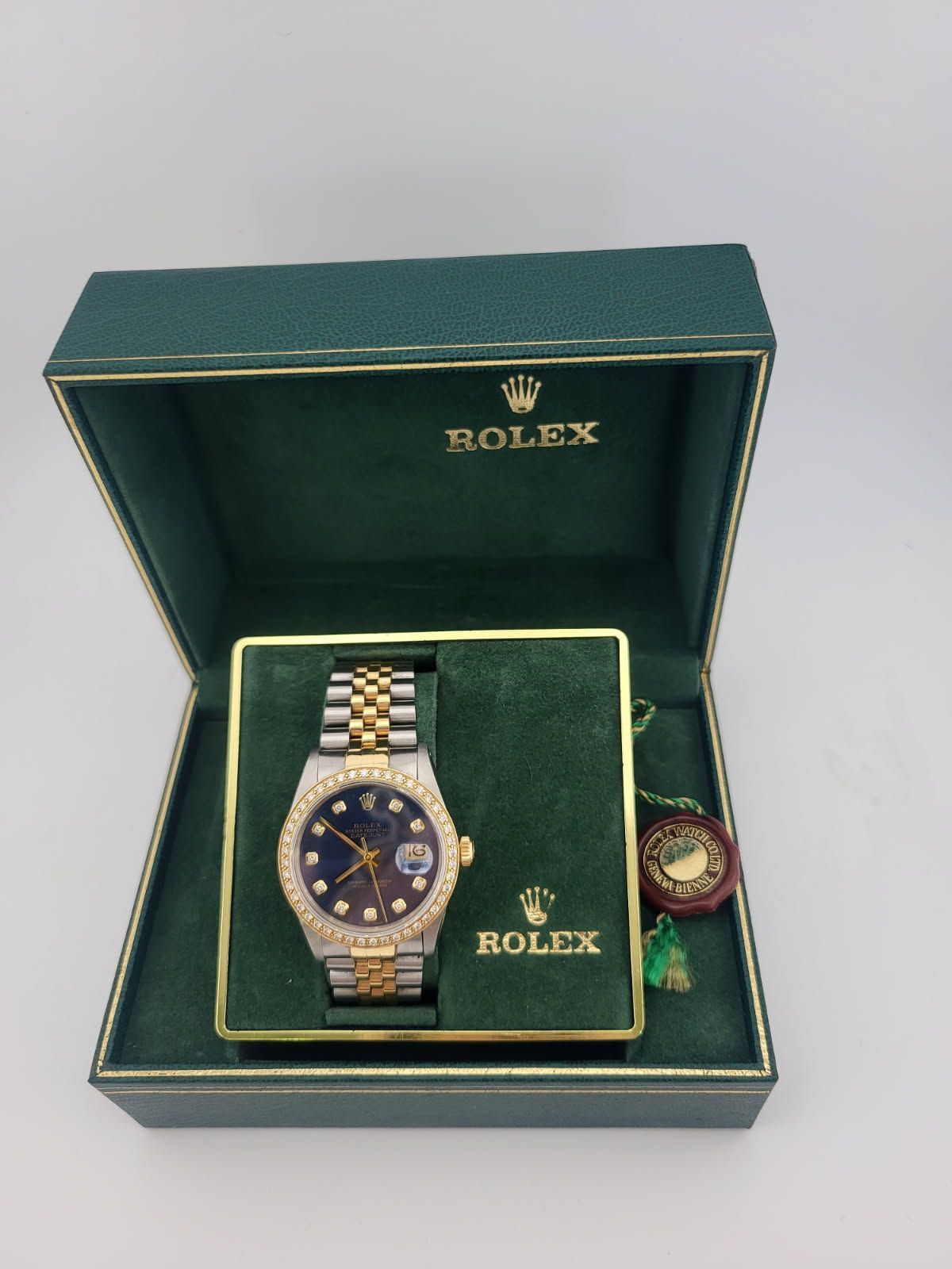 Rolex watch in green box with gold trim. Two-tone metal band, diamond-encrusted bezel, and blue face.