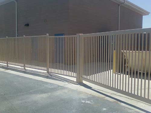 Beige metal fence outside a building, with vertical bars and a gate.
