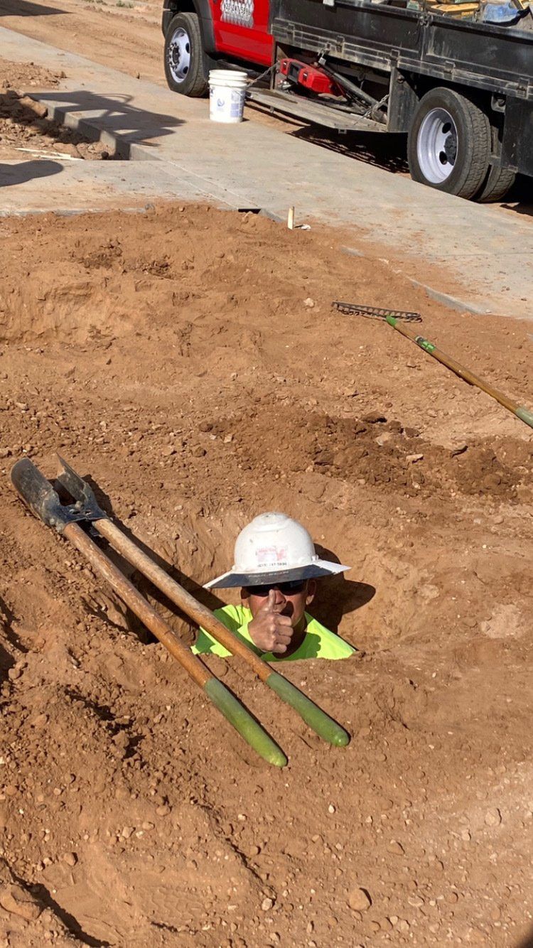 Construction worker in a dirt hole, wearing a white hat. Shovels lie beside him.