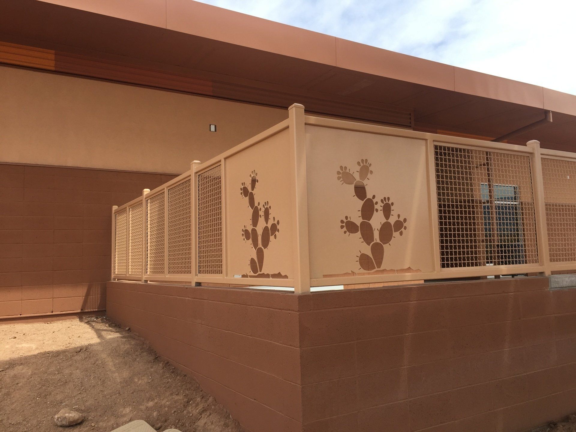 Tan metal fence with cactus cutouts against a brown building, under a blue sky.