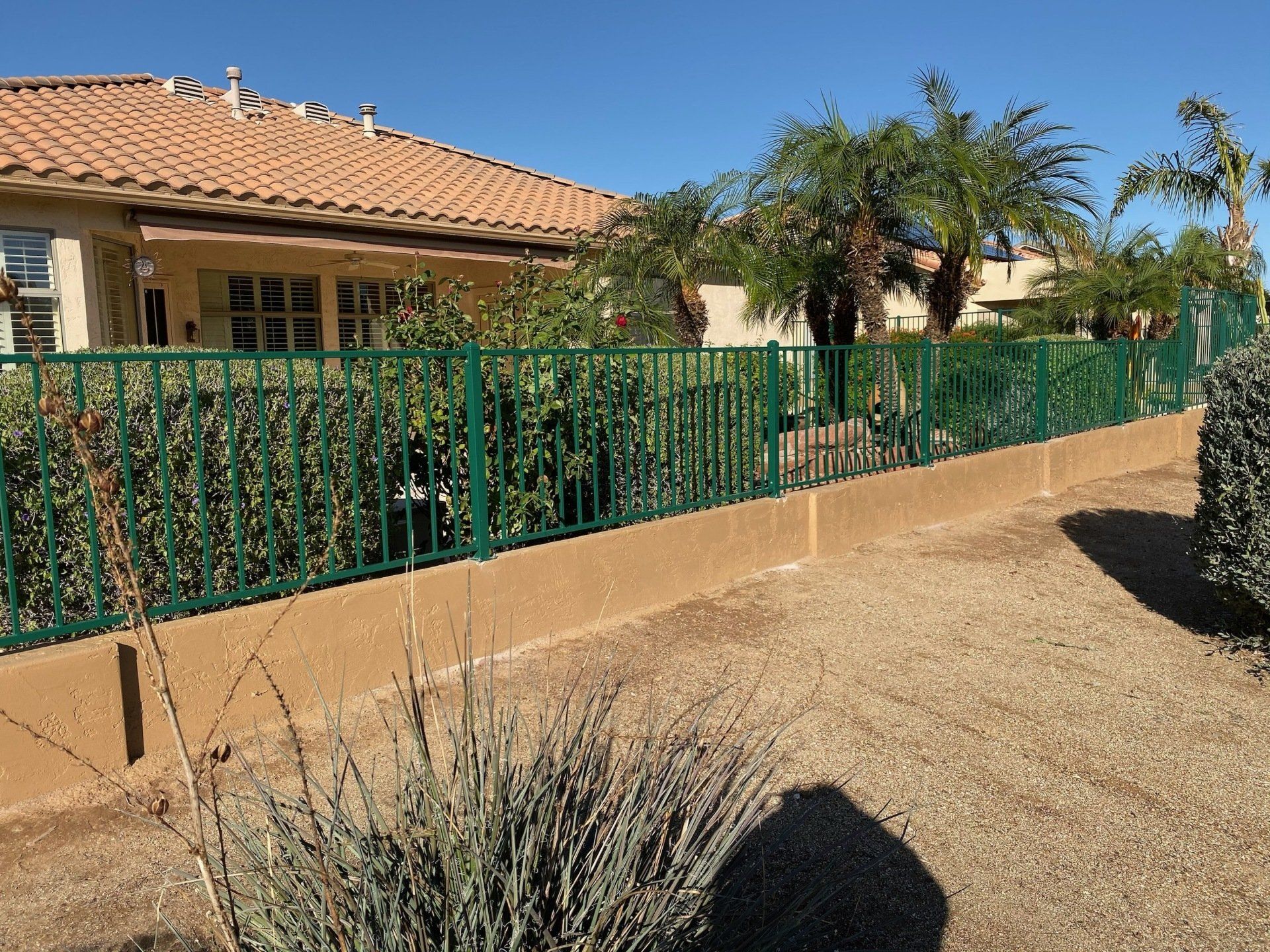 Green metal fence atop a tan wall, separating a gravel yard from a house with a red tile roof and green bushes.