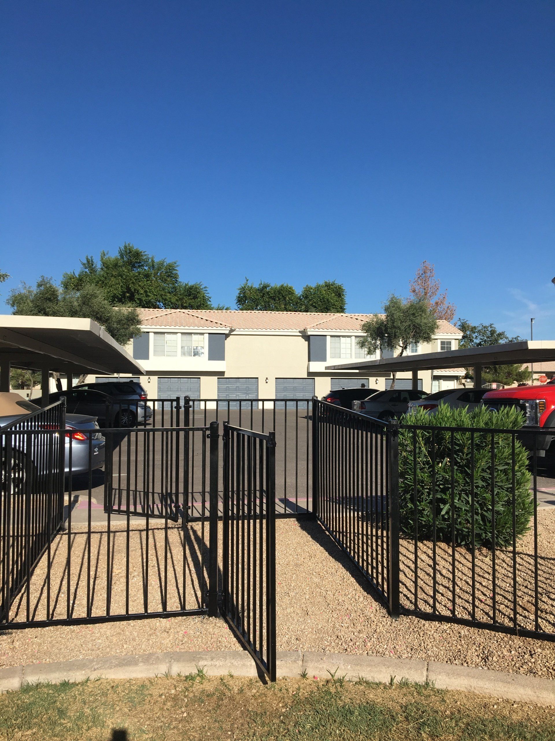 Black gate in front of tan two-story building with carports and cars. Blue sky.