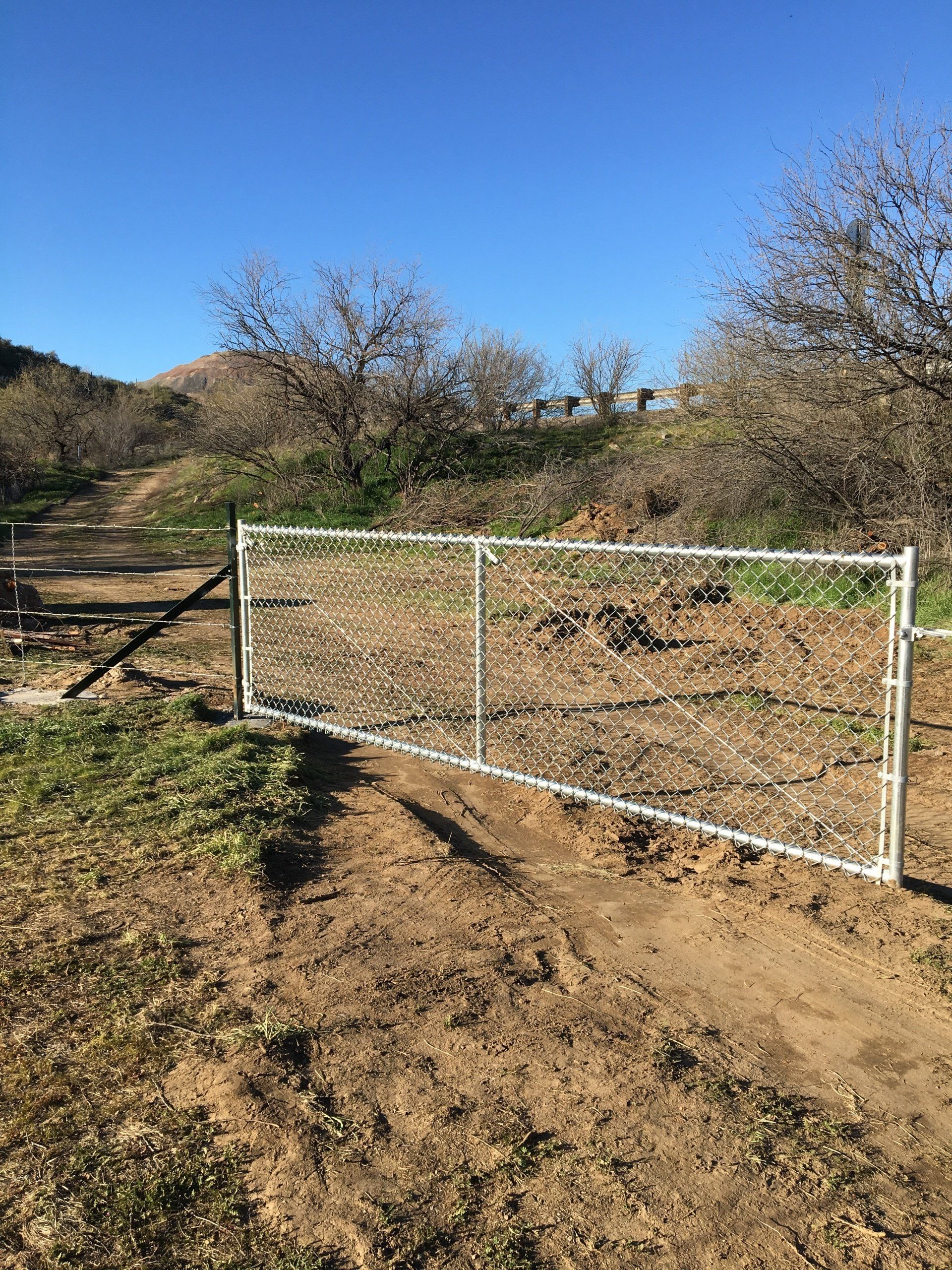 A chain-link gate, white frame, is closed on a dirt path, leading to a hillside under a clear sky.
