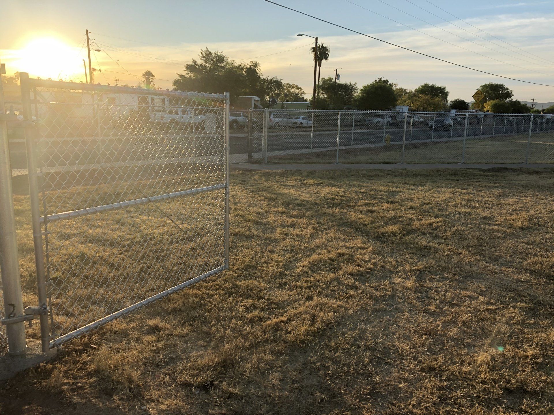 Open chain-link gate in a grassy field at sunset. Fenced area with buildings and trees in the background.