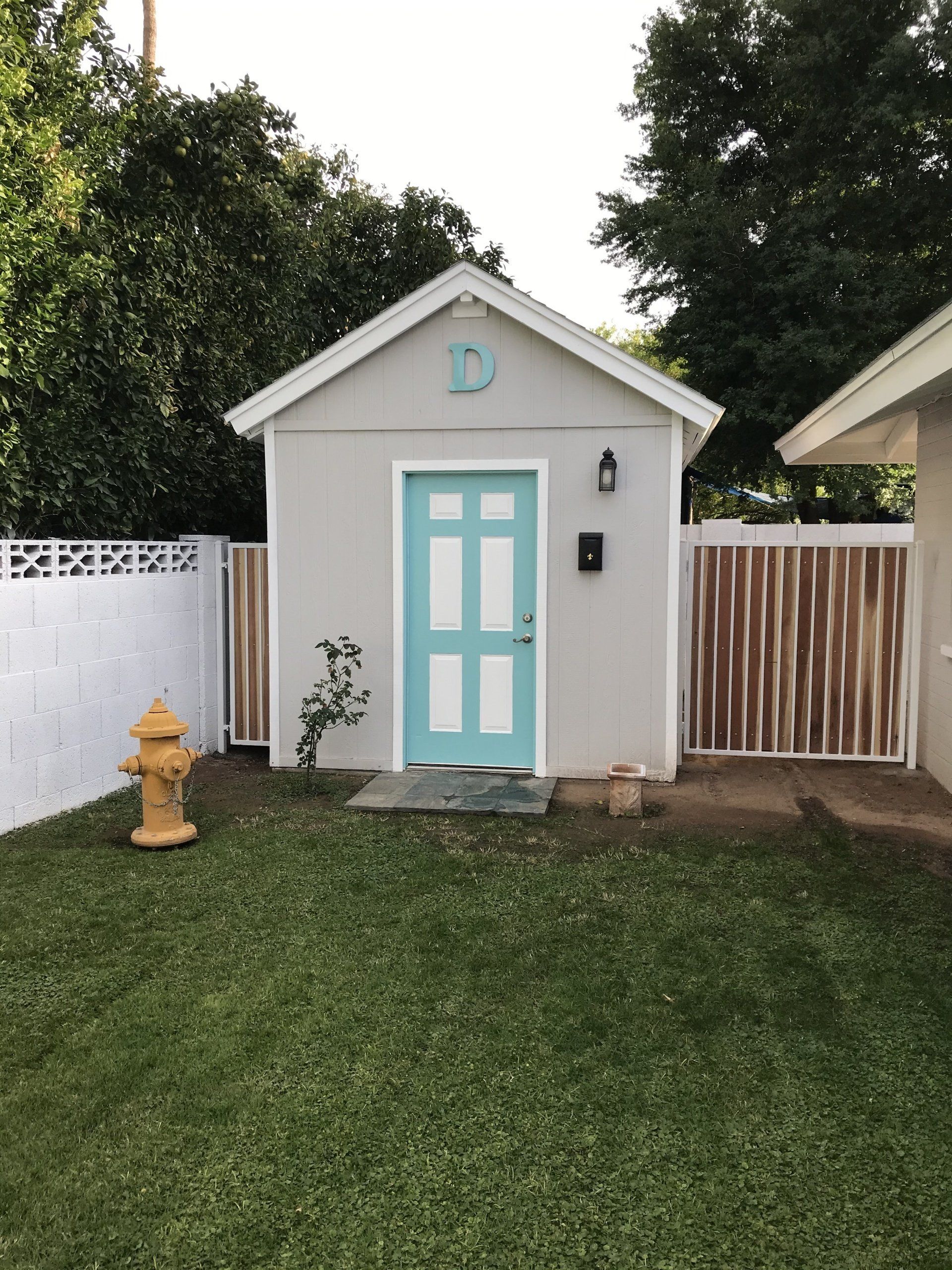 Small gray shed with a teal door, yellow fire hydrant, and white and corrugated fences in a grassy yard.