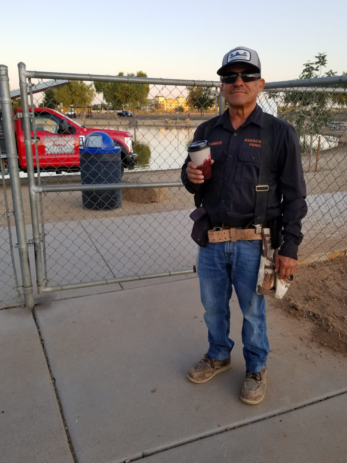 Man in work clothes holding coffee, standing by a fence, a red truck, and a body of water in the background.