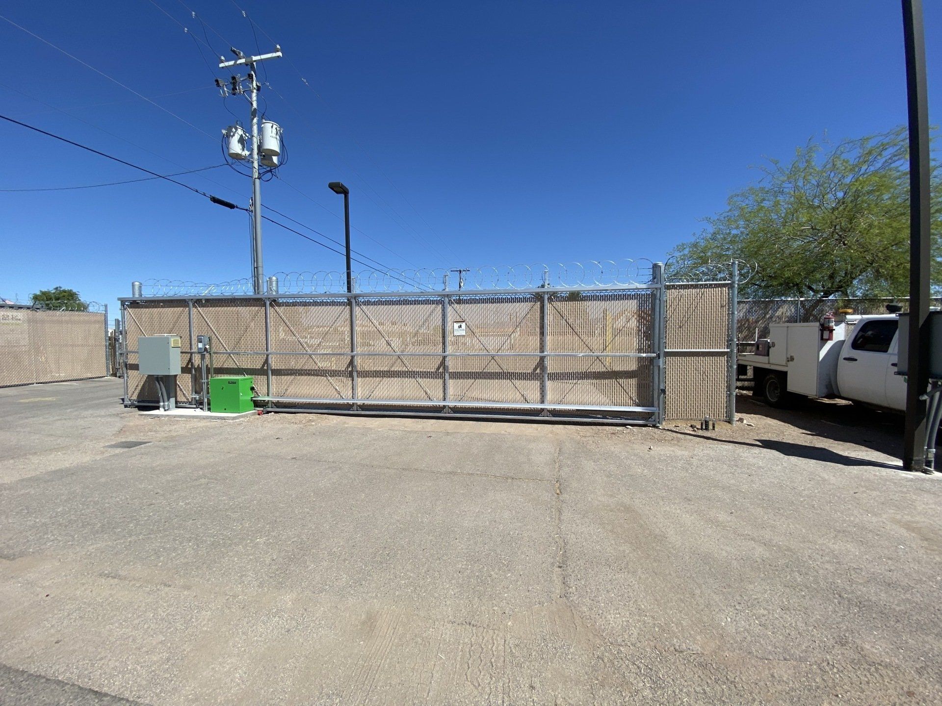 A large, closed metal gate with barbed wire atop. Gray gravel driveway, blue sky. A power pole and white truck visible.
