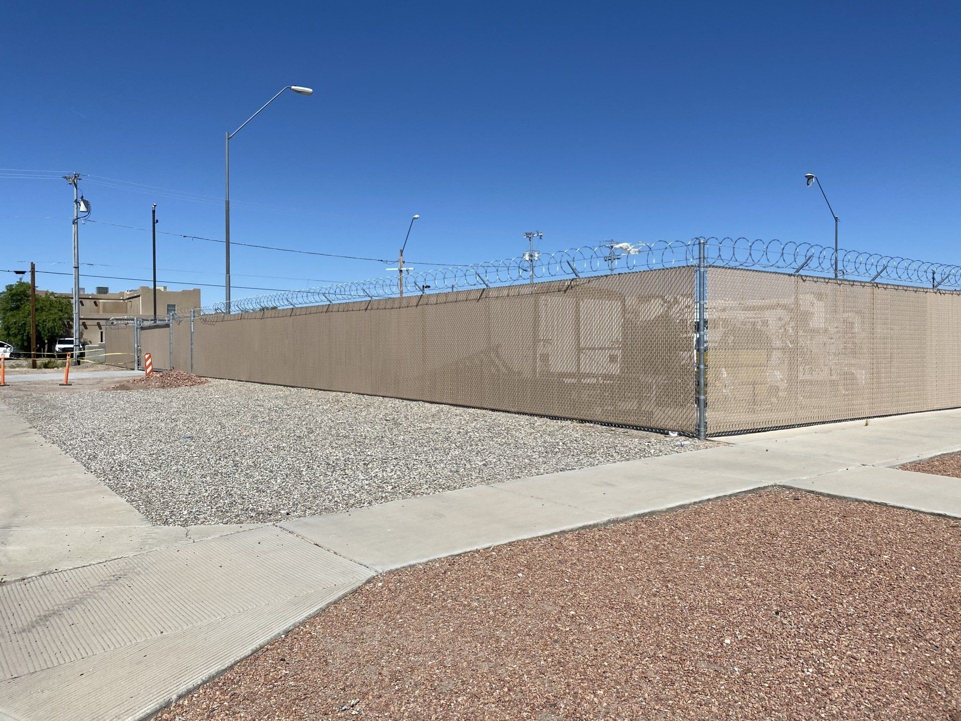 A beige chain-link fence with barbed wire tops encloses a facility, set against a bright blue sky.
