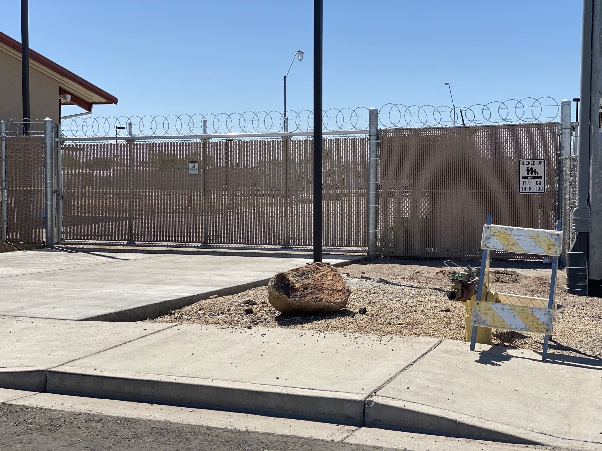 A gated entrance with barbed wire on top. A large rock sits in front, and a barrier is on the right.
