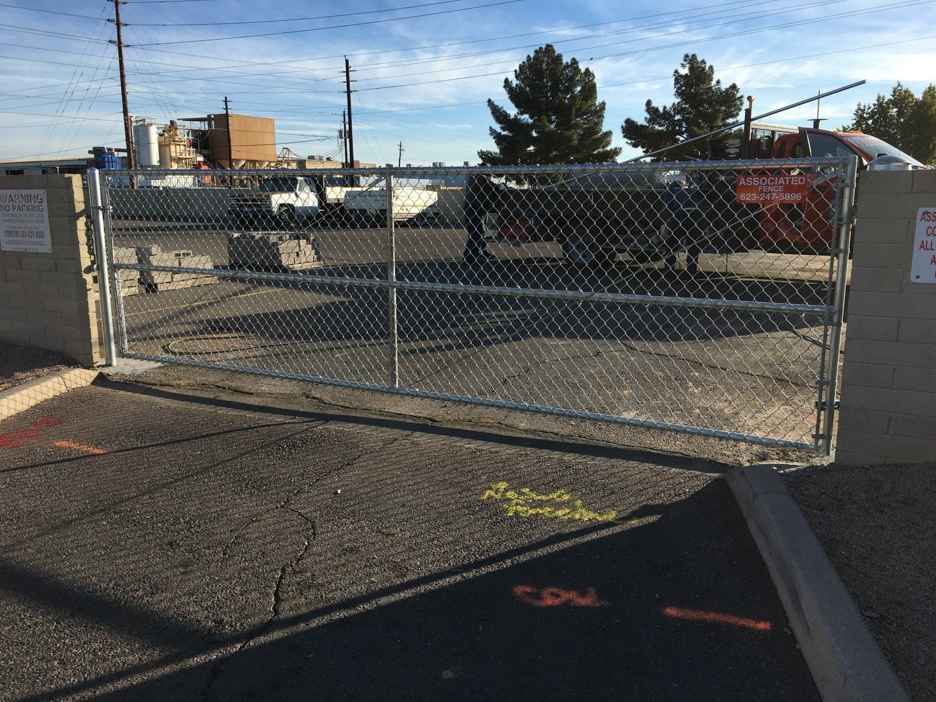 Chain-link gate in front of a paved area; behind it is a work site with vehicles and buildings.