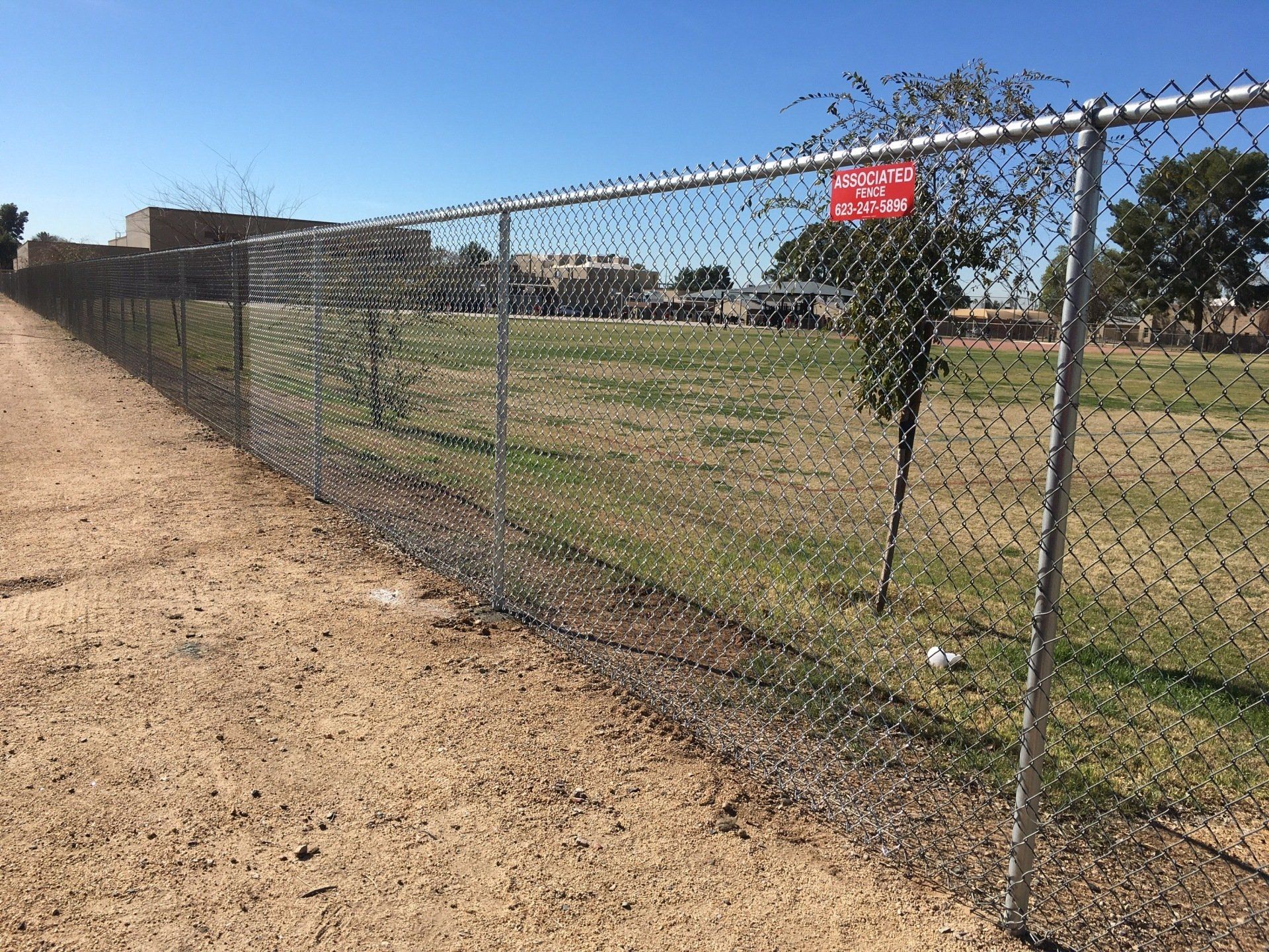 Chain-link fence bordering a grassy field and dirt path on a sunny day.