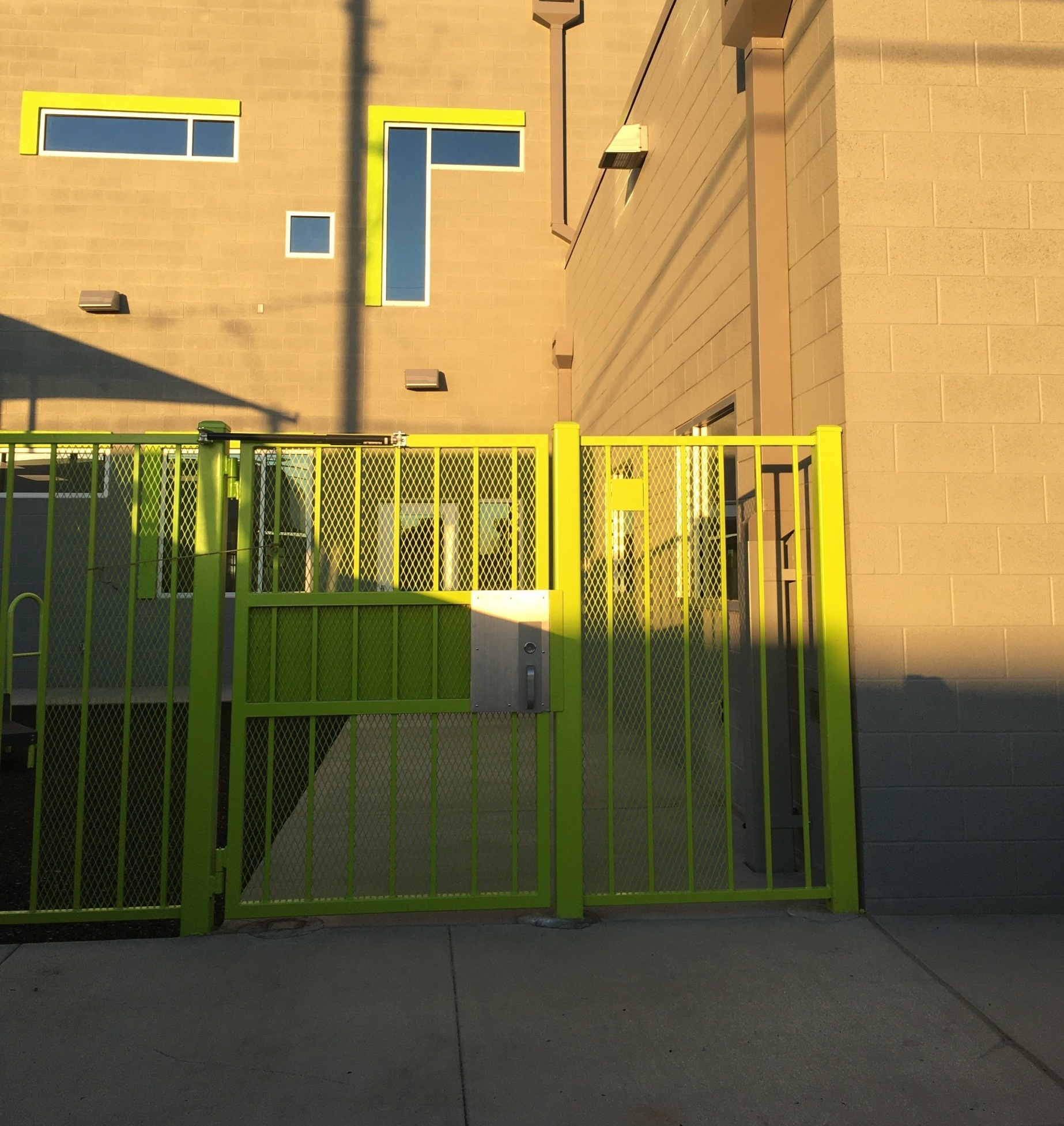 Green gate in front of a building with yellow-trimmed windows and a tan facade, lit by late afternoon sunlight.