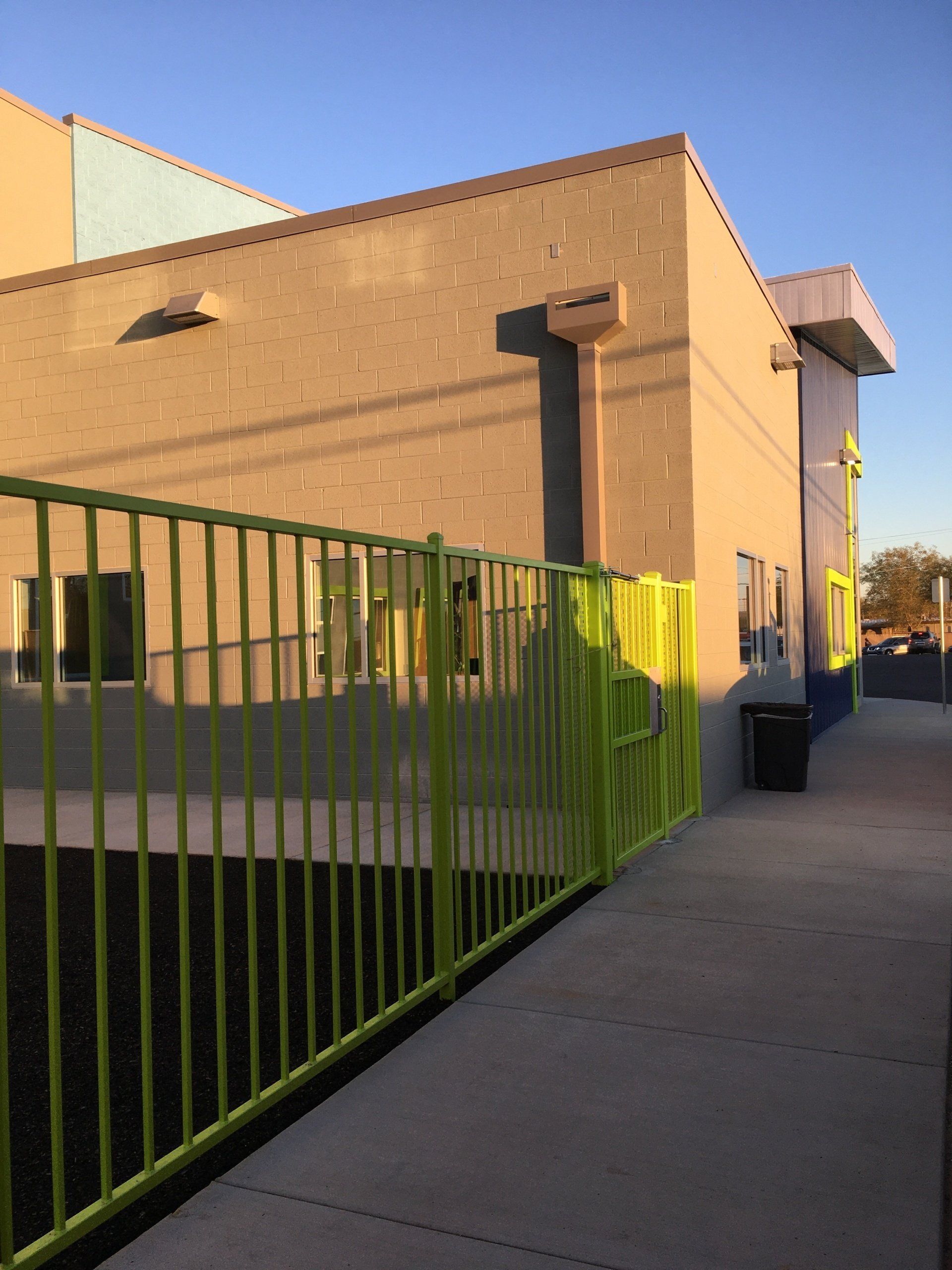 Exterior of a building with a green fence and sidewalk. Sunny day with shadows.