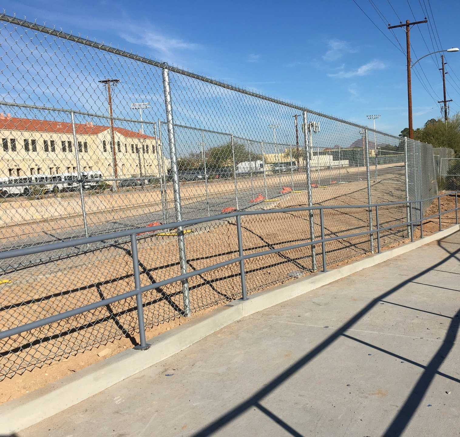 Chain-link fence with handrail beside a sidewalk. Background: building, power lines, and clear sky.