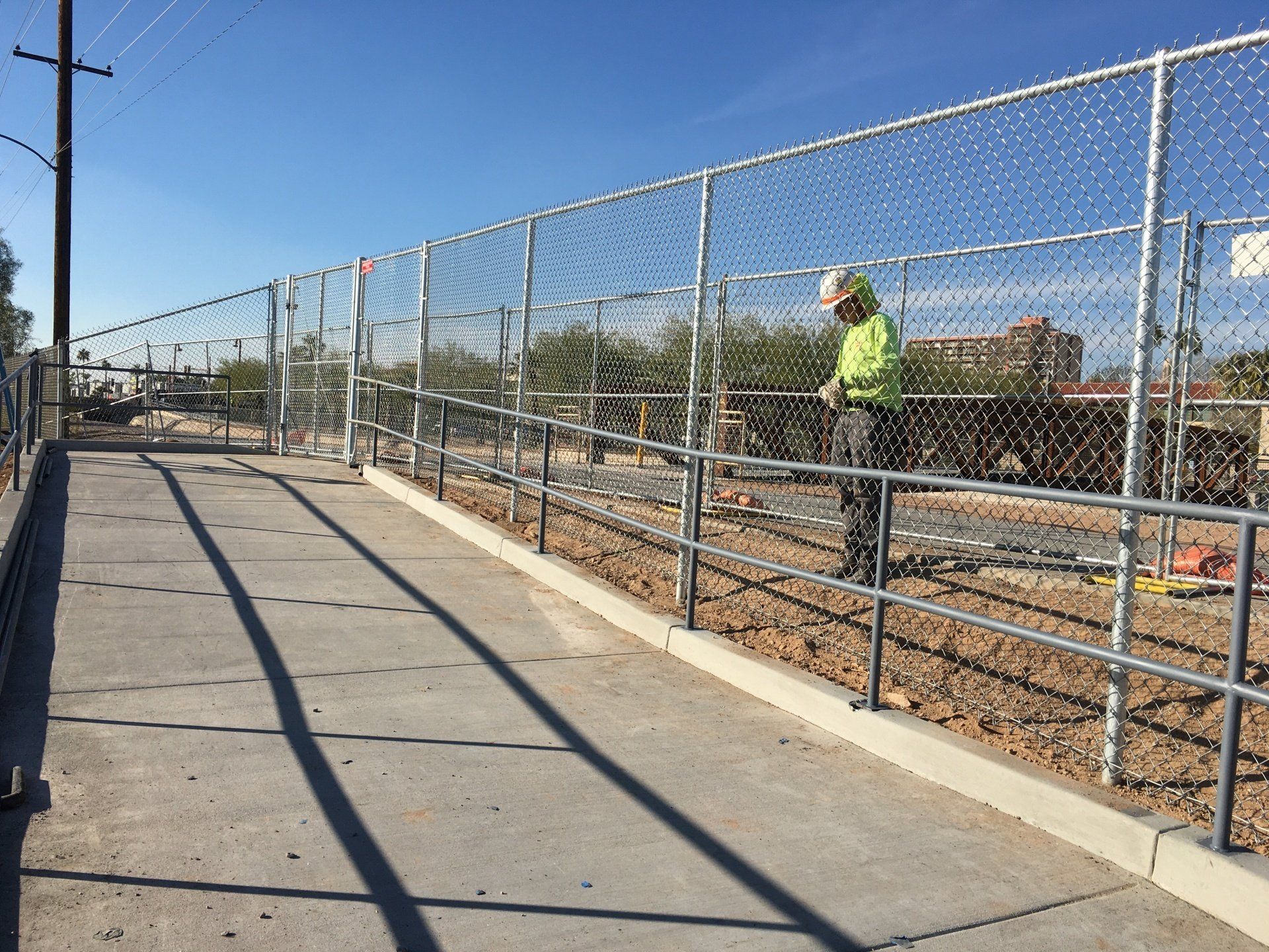 Worker in safety vest near chain-link fence and concrete walkway, outdoors on a sunny day.