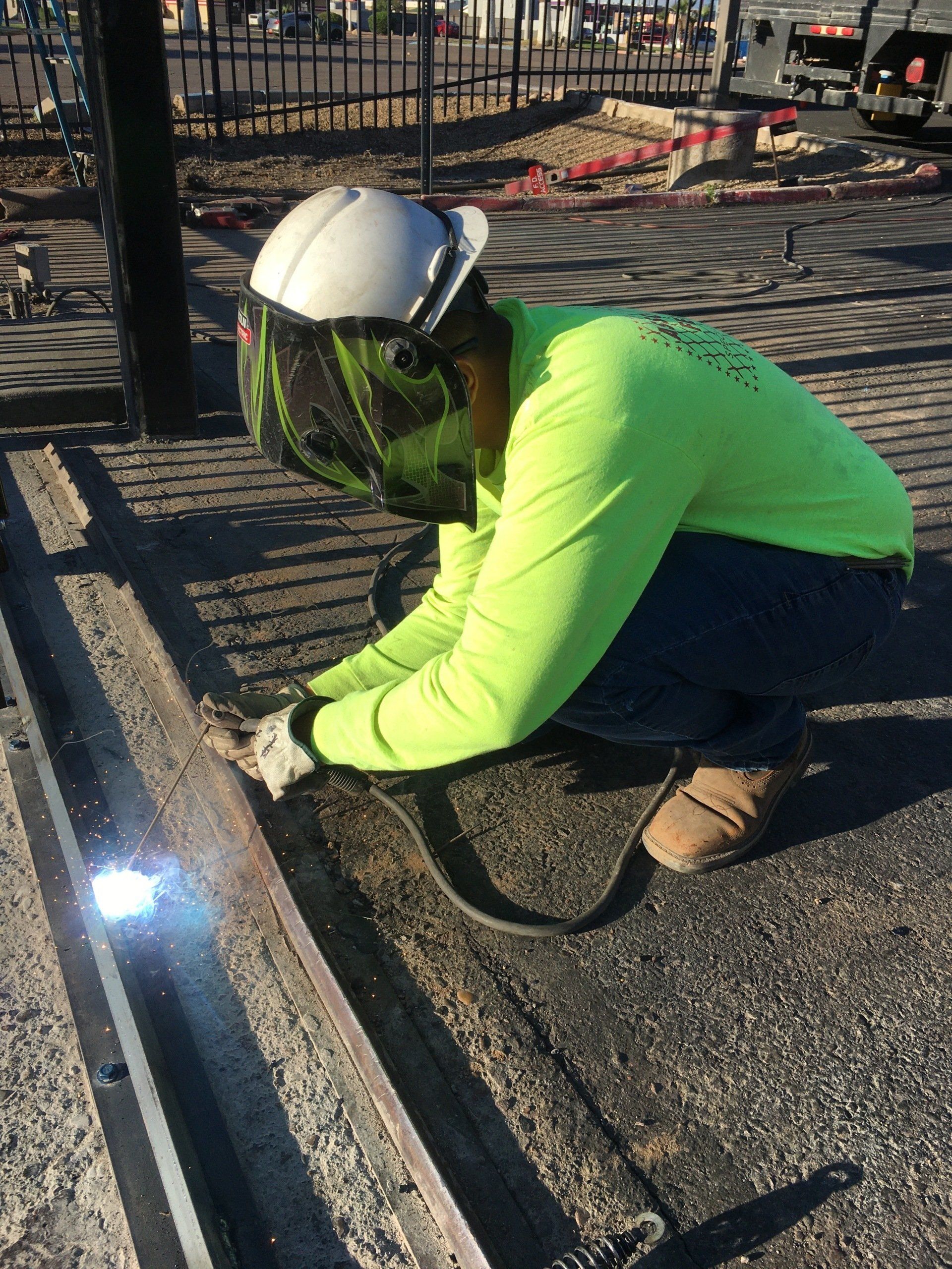 Welder in neon green shirt, protective helmet, welding metal on pavement.