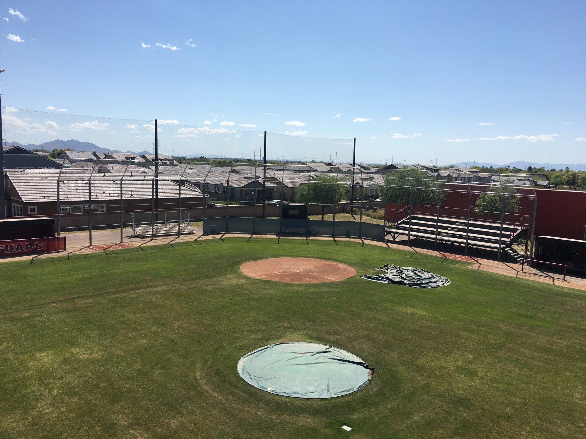 Baseball field with green grass, brown dirt, and a blue sky. Fences surround the field, with houses in the background.