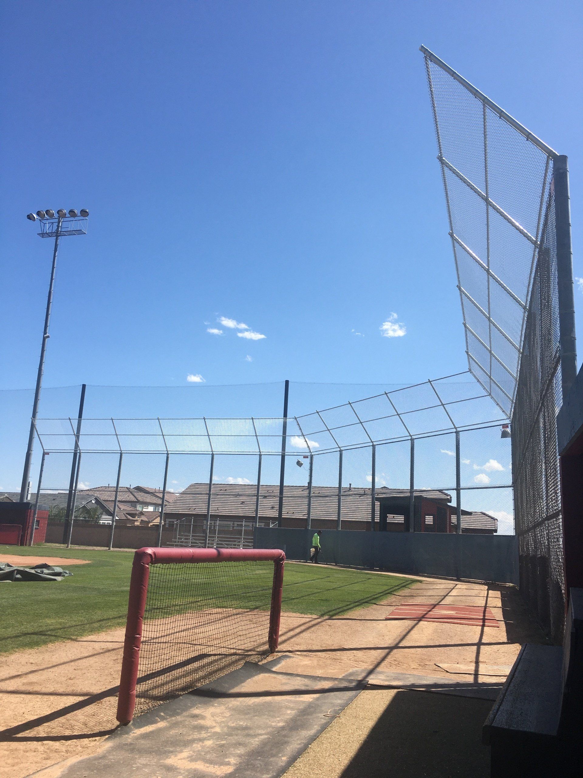 Baseball field with tall netting, blue sky. A red gate and light pole are visible.