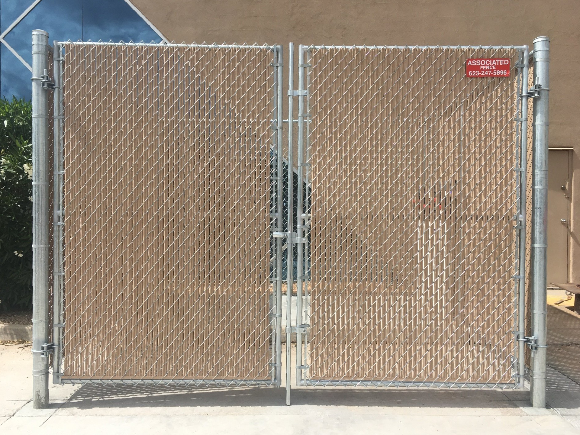 Double chain-link gate with tan privacy slats, silver frame, concrete ground, and tan building background.
