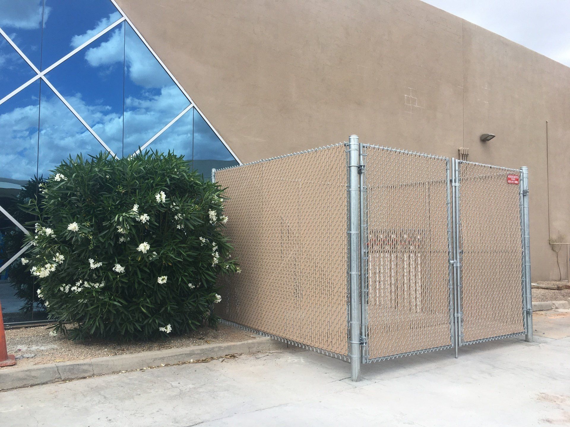 Tan chain-link fence with tan privacy slats beside a beige building and blue glass. A green bush is beside the fence.