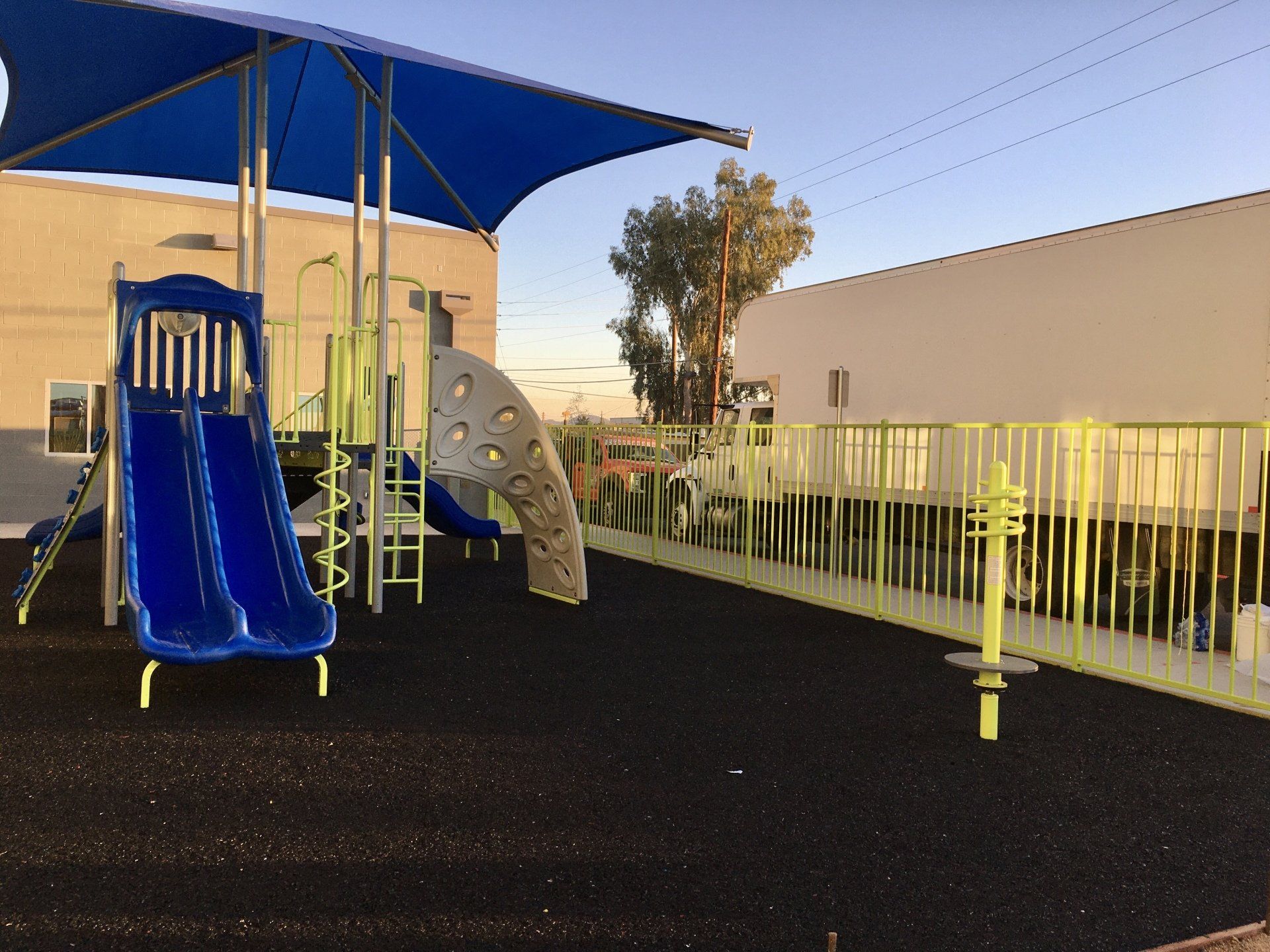 Playground with blue slides, climbing wall, and shade cover. Black rubber ground and yellow fence.