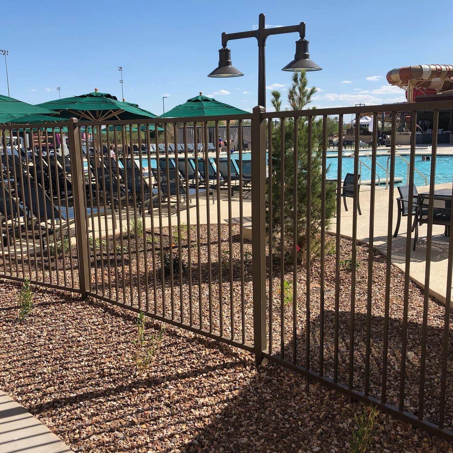 Brown metal fence bordering a pool area with lounge chairs, umbrellas, and landscaping under a sunny sky.