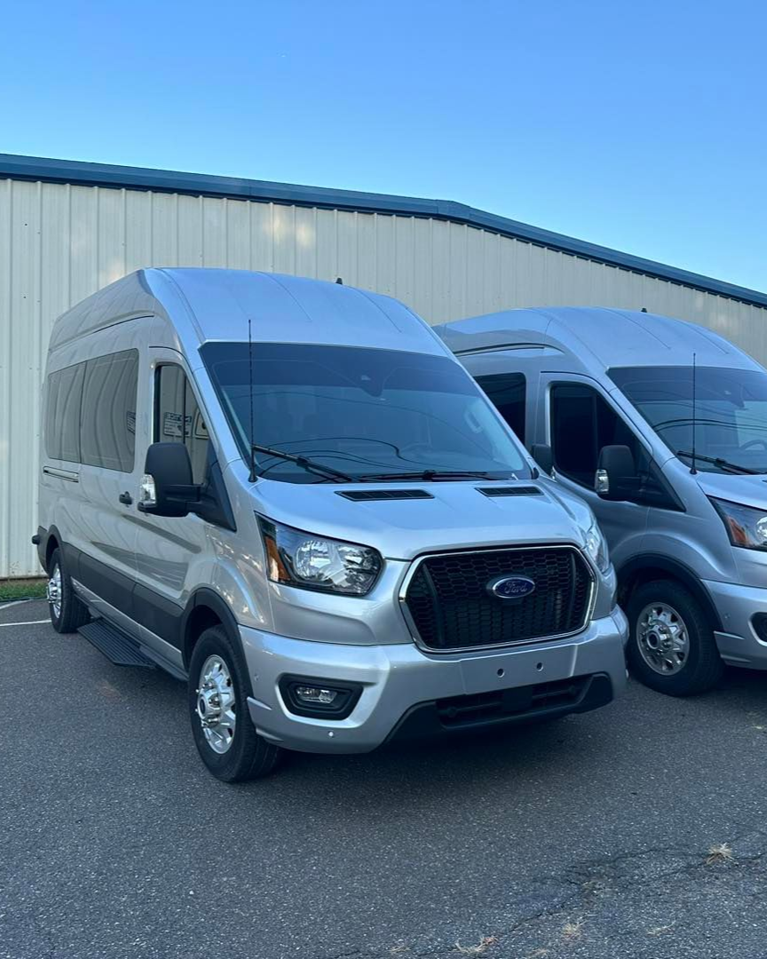 Two silver Ford Transit passenger vans parked outdoors next to a building wall.