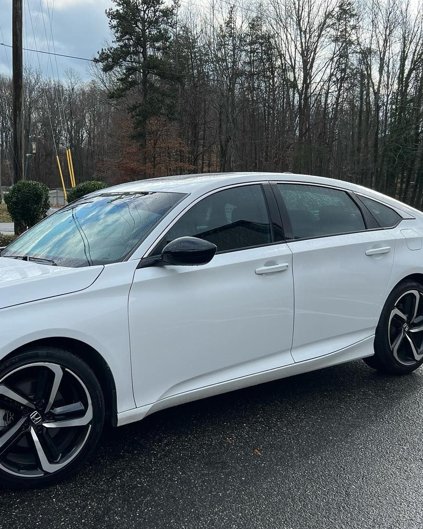 A white sedan parked on a paved surface with a wooded area in the background.