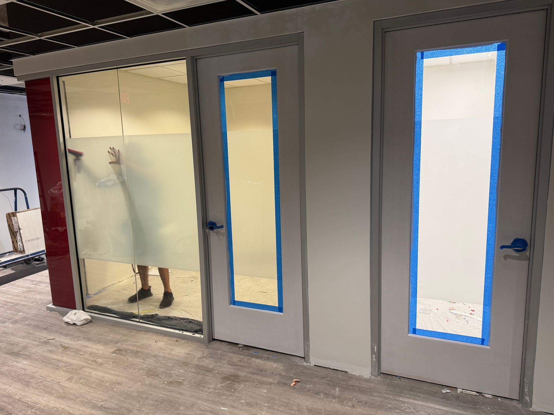 A person applies frosted window film to a glass office wall next to two doors with blue painter's tape on their windows.