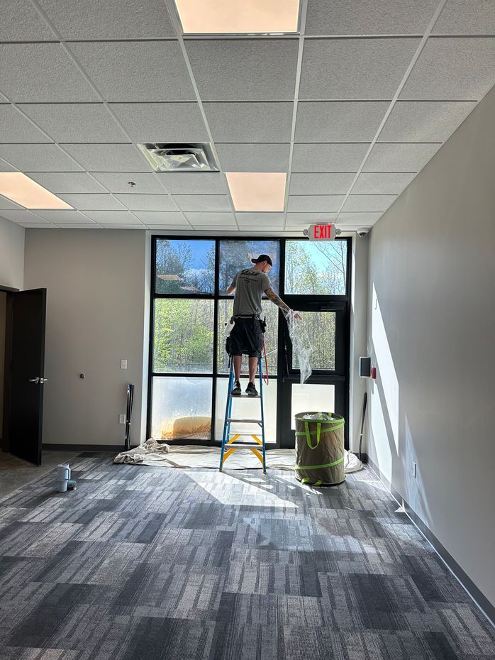 A person stands on a step ladder applying film to a large office window in a room with gray carpet and a drop ceiling.