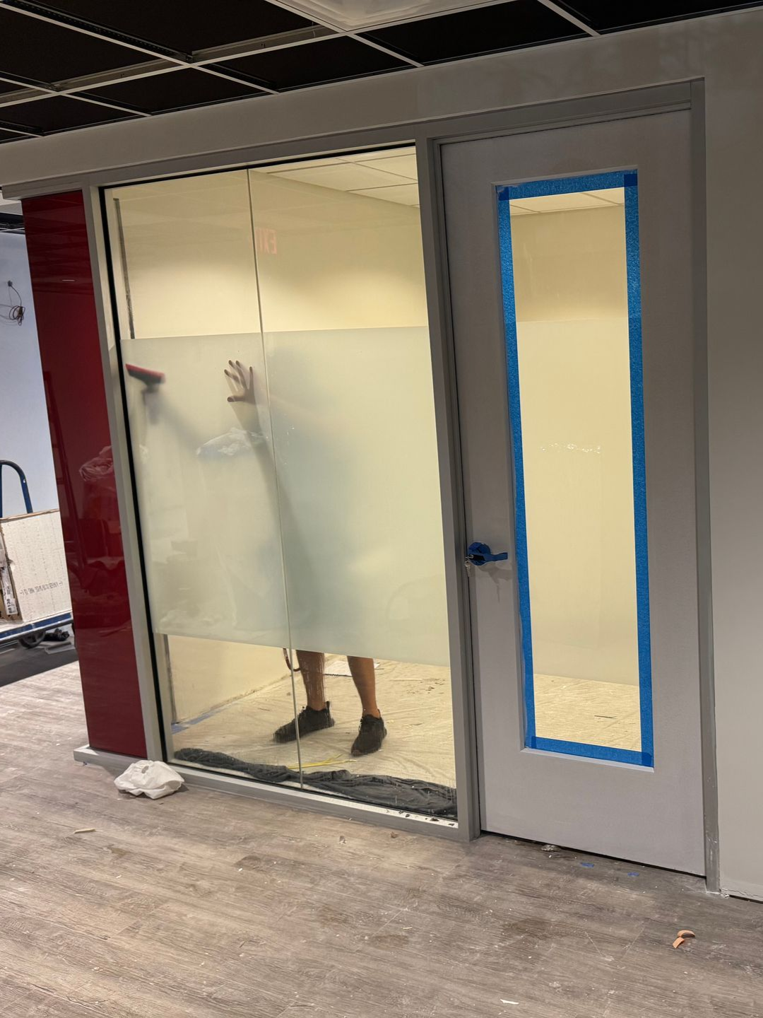 A person applying frosted privacy film to a large glass office partition beside a doorway with blue painter's tape.