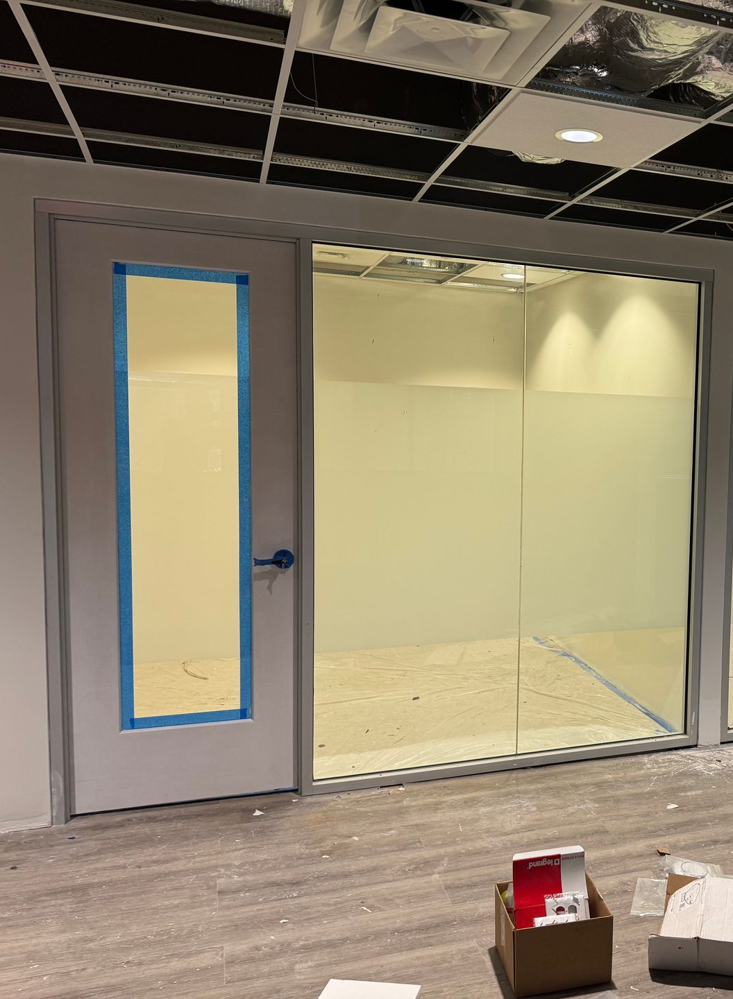 A person on a ladder cleans a large window in a gray-toned, empty office space with a patterned carpet.