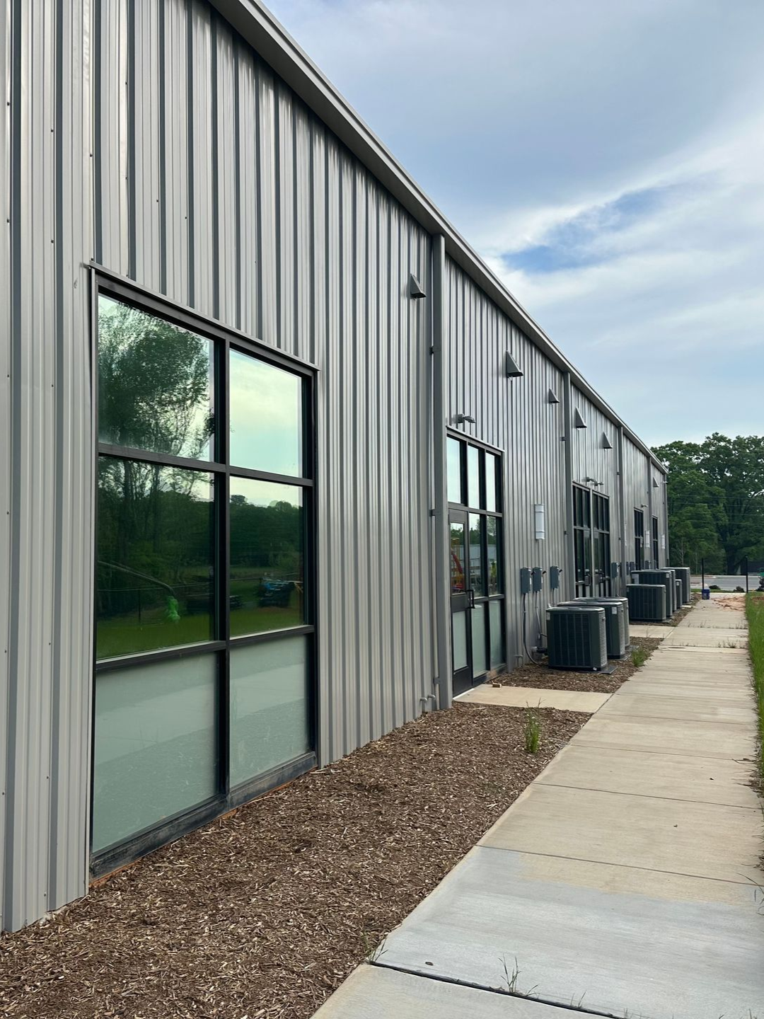Exterior view of a long, silver metal industrial building with windows, a concrete walkway, and outdoor AC units.