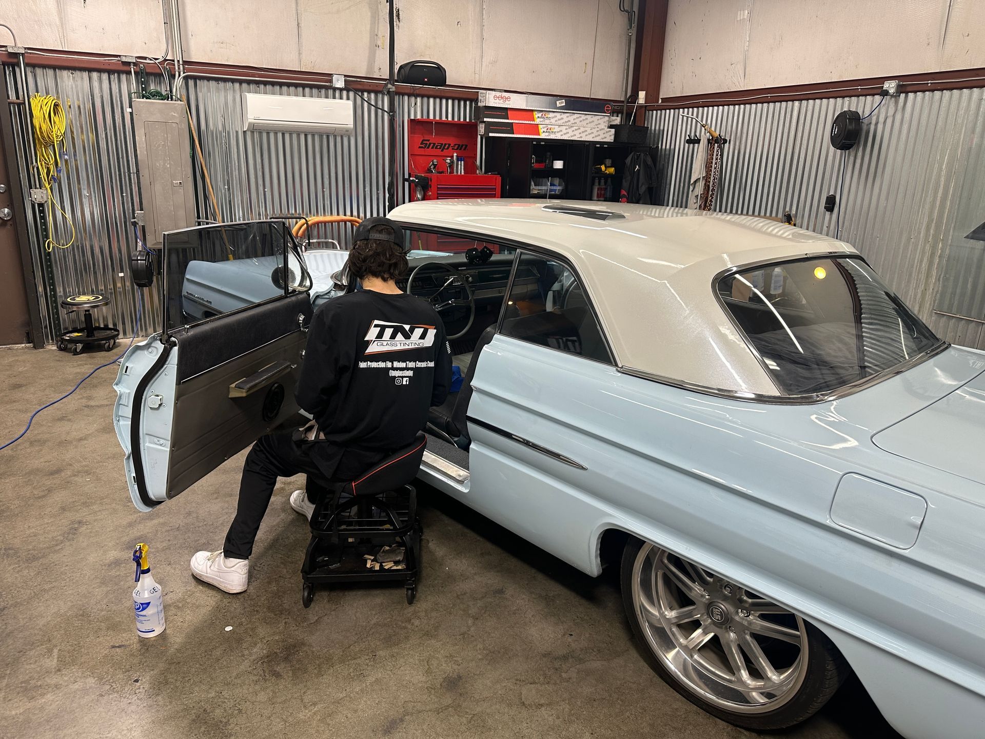 A person in a black sweatshirt sits on a rolling stool, working on the interior of a light blue and white vintage car.