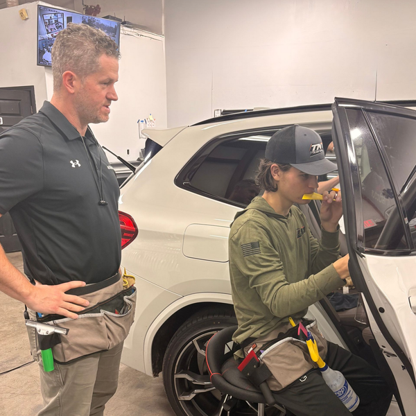 Two people in a garage focus on working on the interior of a white car, one standing and one seated with a tool.