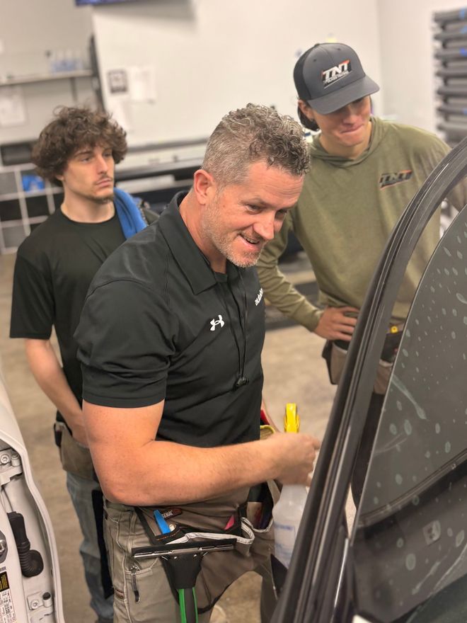 A man in a black polo shirt holds a spray bottle while instructing two observers in a workshop setting.