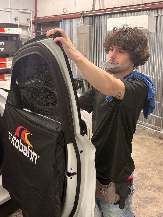 A person with curly hair wearing a black shirt works on a vehicle door cover with a Autobahn logo in a workshop.