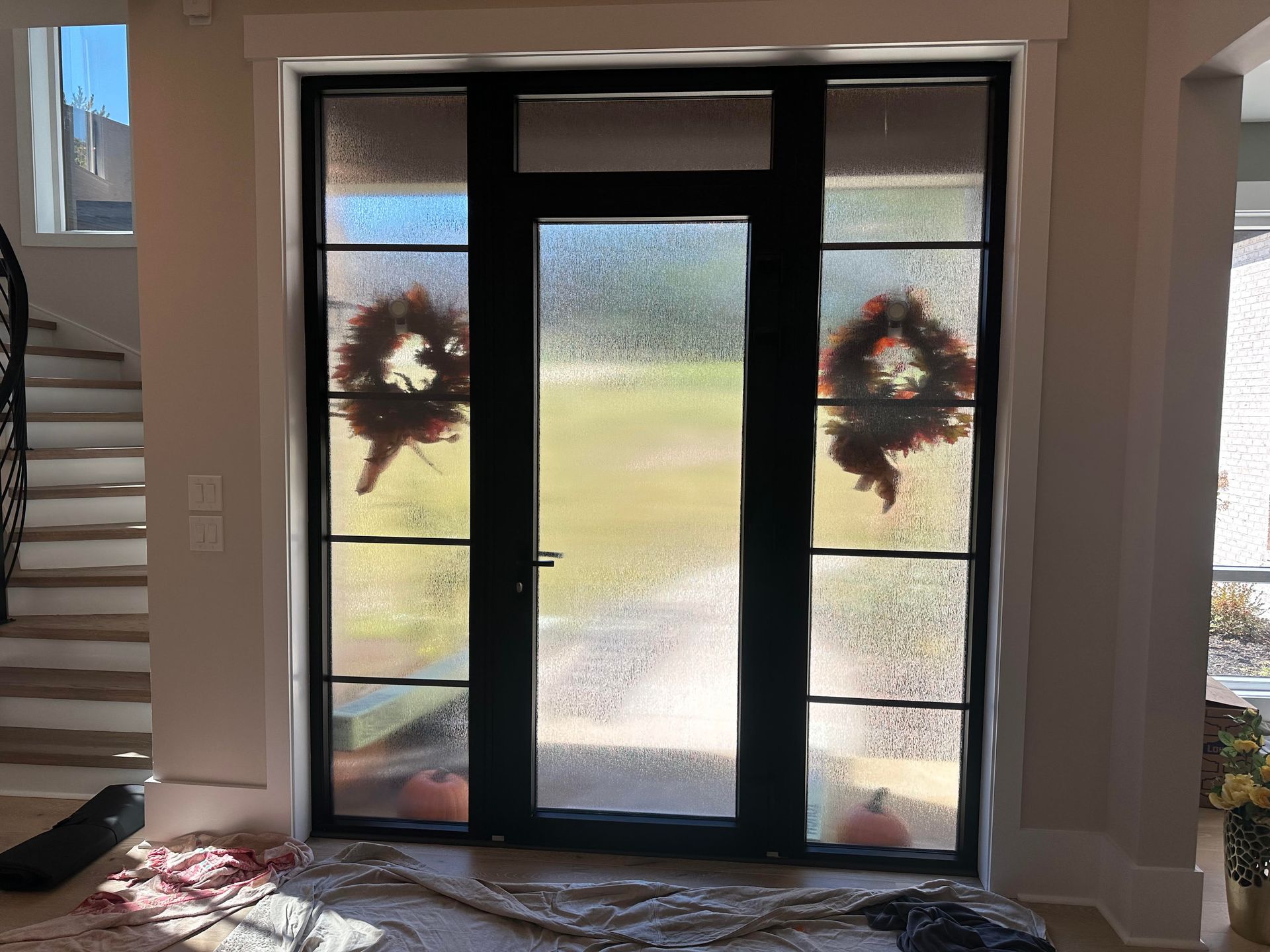 A black-framed glass front door with textured panels and side windows, each featuring a decorative autumn wreath.